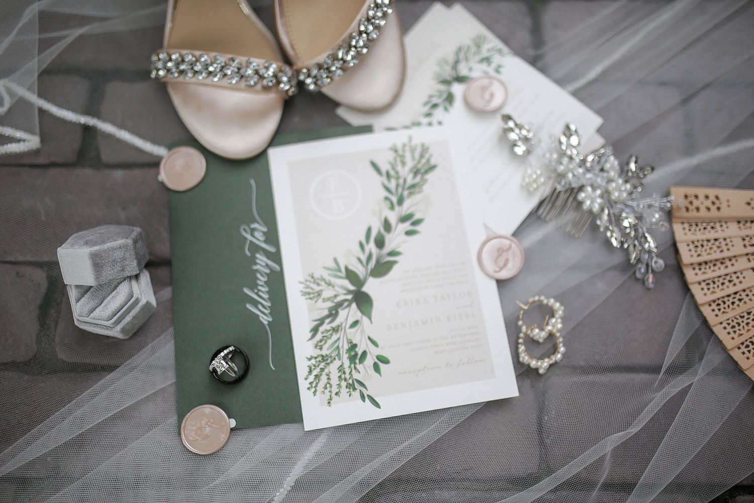Wedding invitation suite with greenery details and jewelry on gray background.