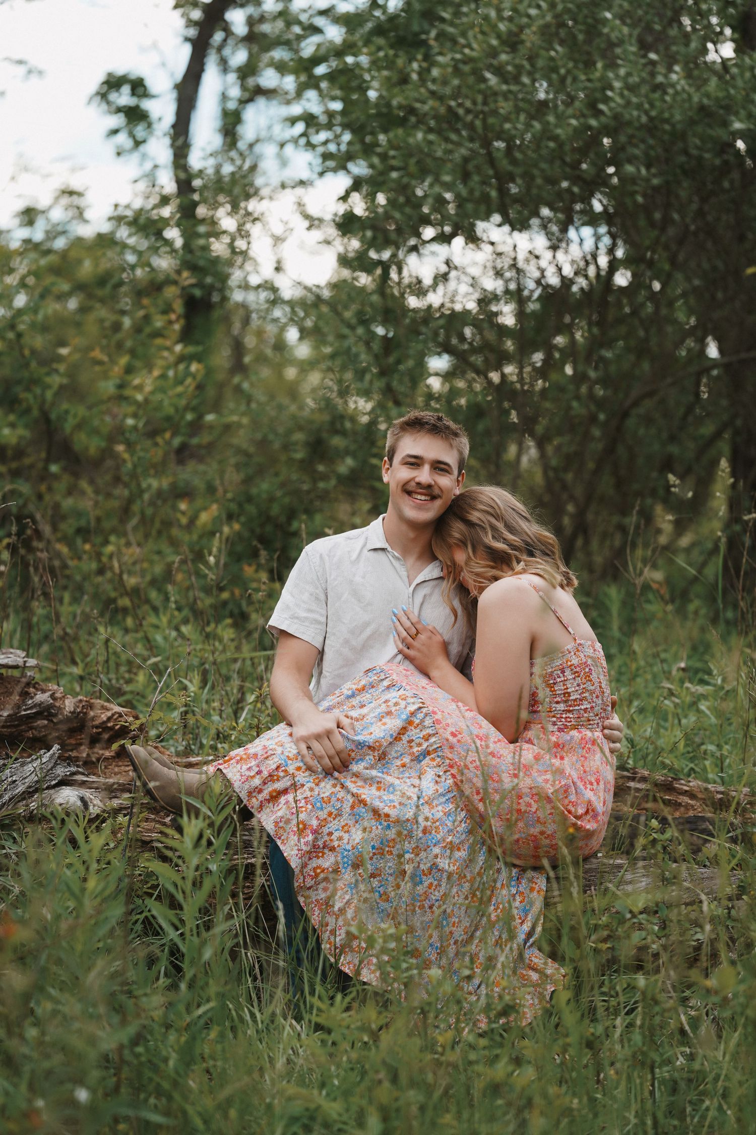 Couple sharing romantic moment while sitting outdoors on a fallen log surrounded by lush greenery.