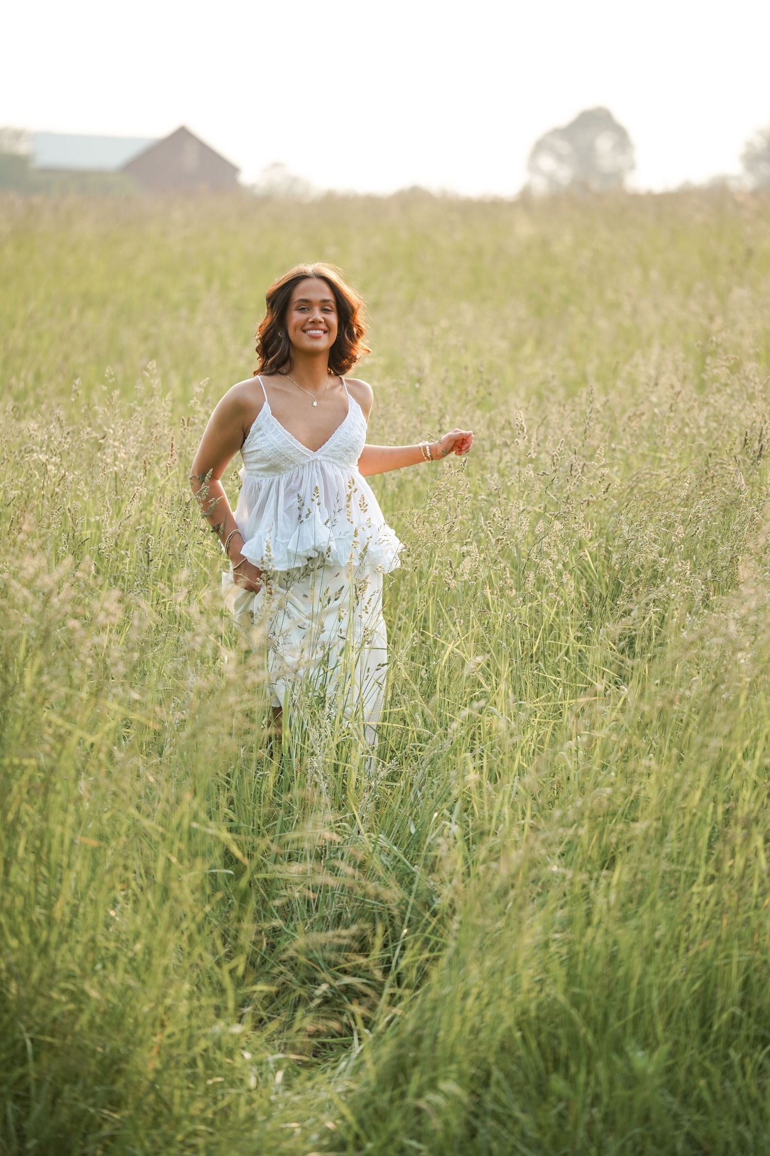 Person in white dress dances joyfully through a golden field of tall grass at sunset.