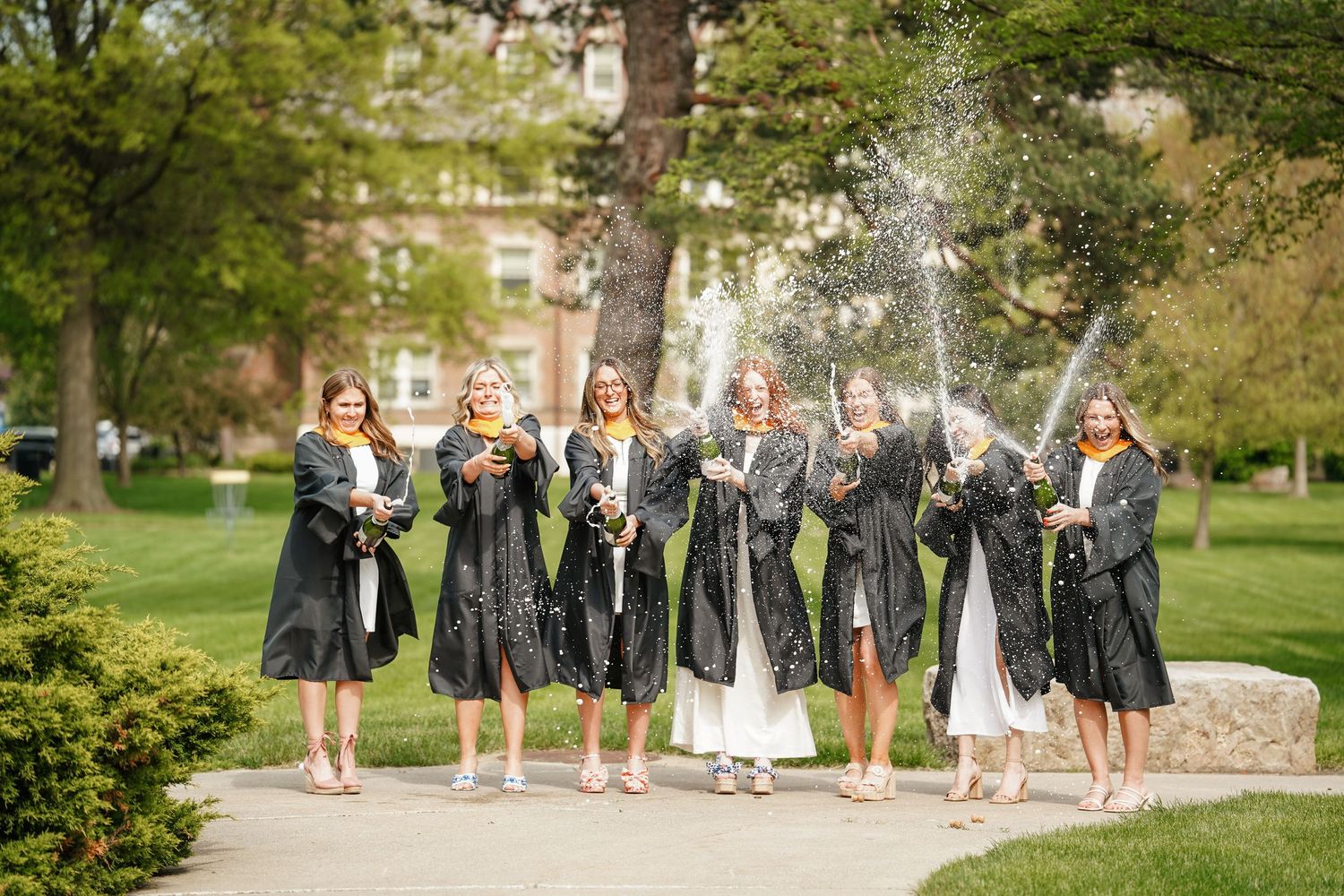 Wedding party walks through a lush garden pathway while holding bouquets and wearing coordinated gray dresses.