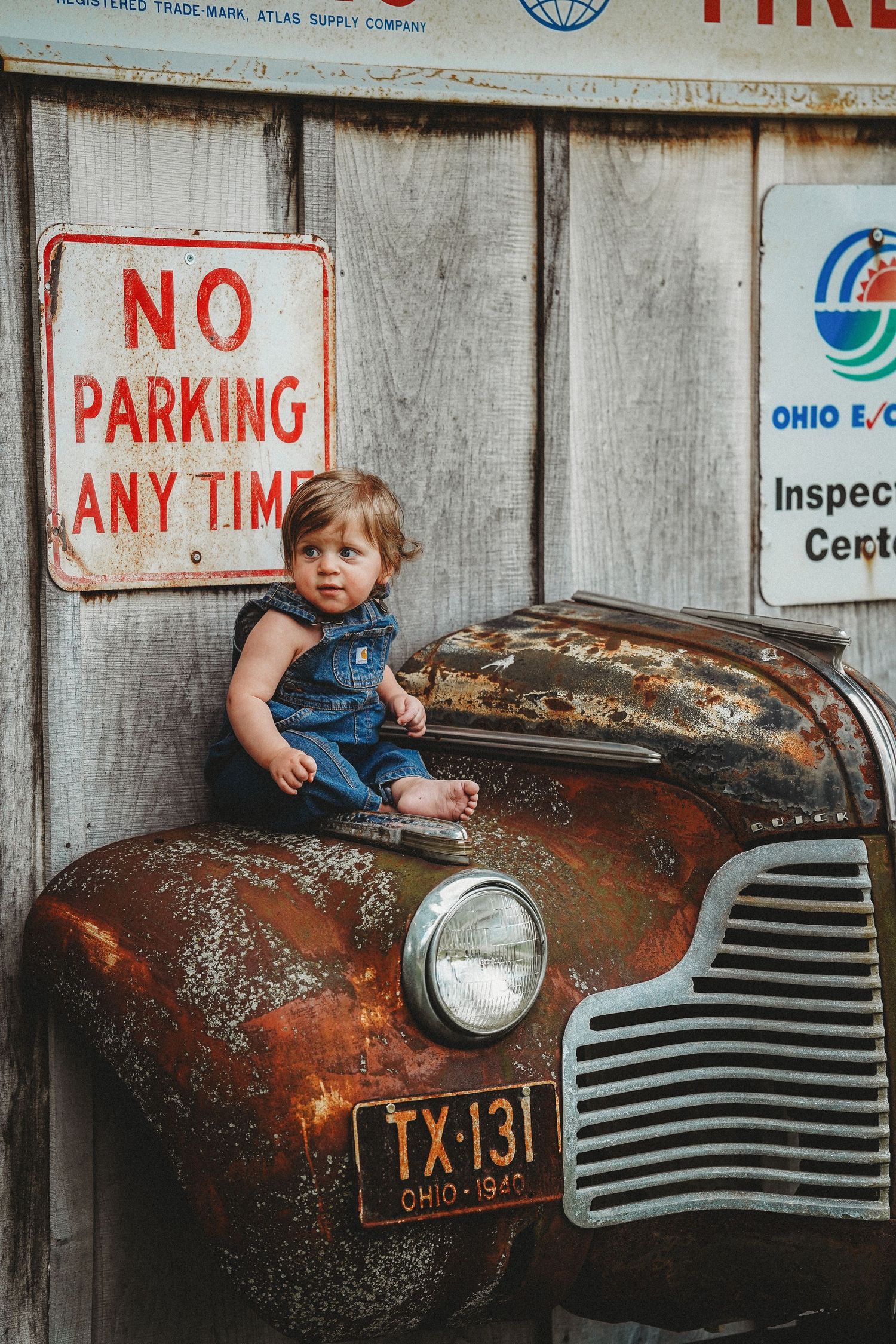 Small child in denim sits on rusty vintage car with Ohio license plate near No Parking sign.