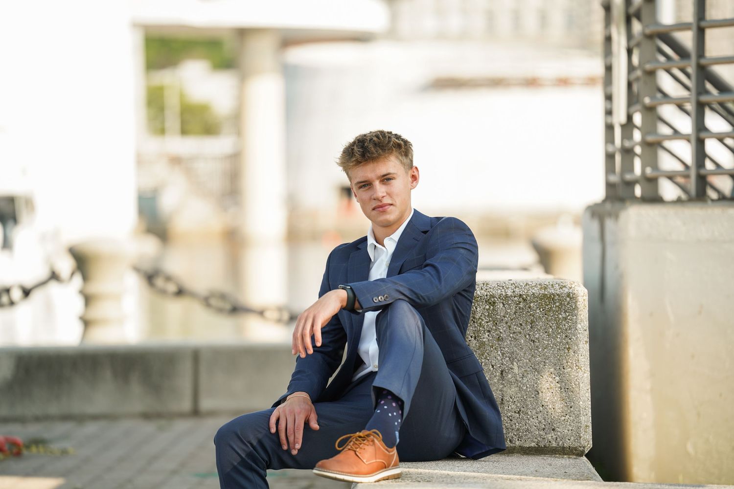 Young man in navy suit and brown dress shoes sits casually on concrete steps outdoors during portrait session.