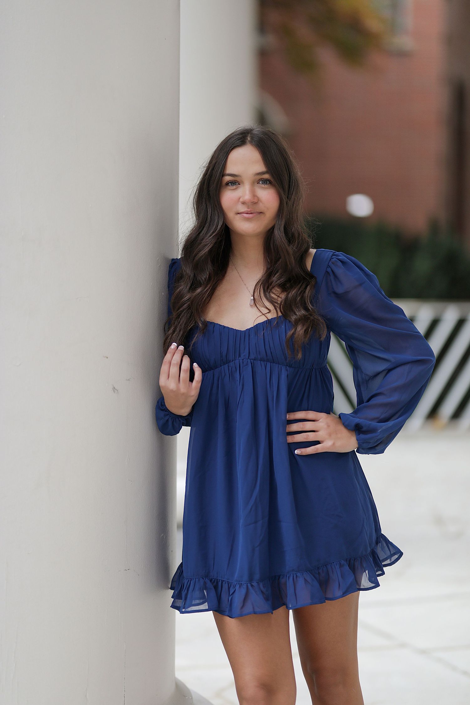 Model wearing a flowy navy blue mini dress with long sleeves and a ruffled hem poses against a white column backdrop.