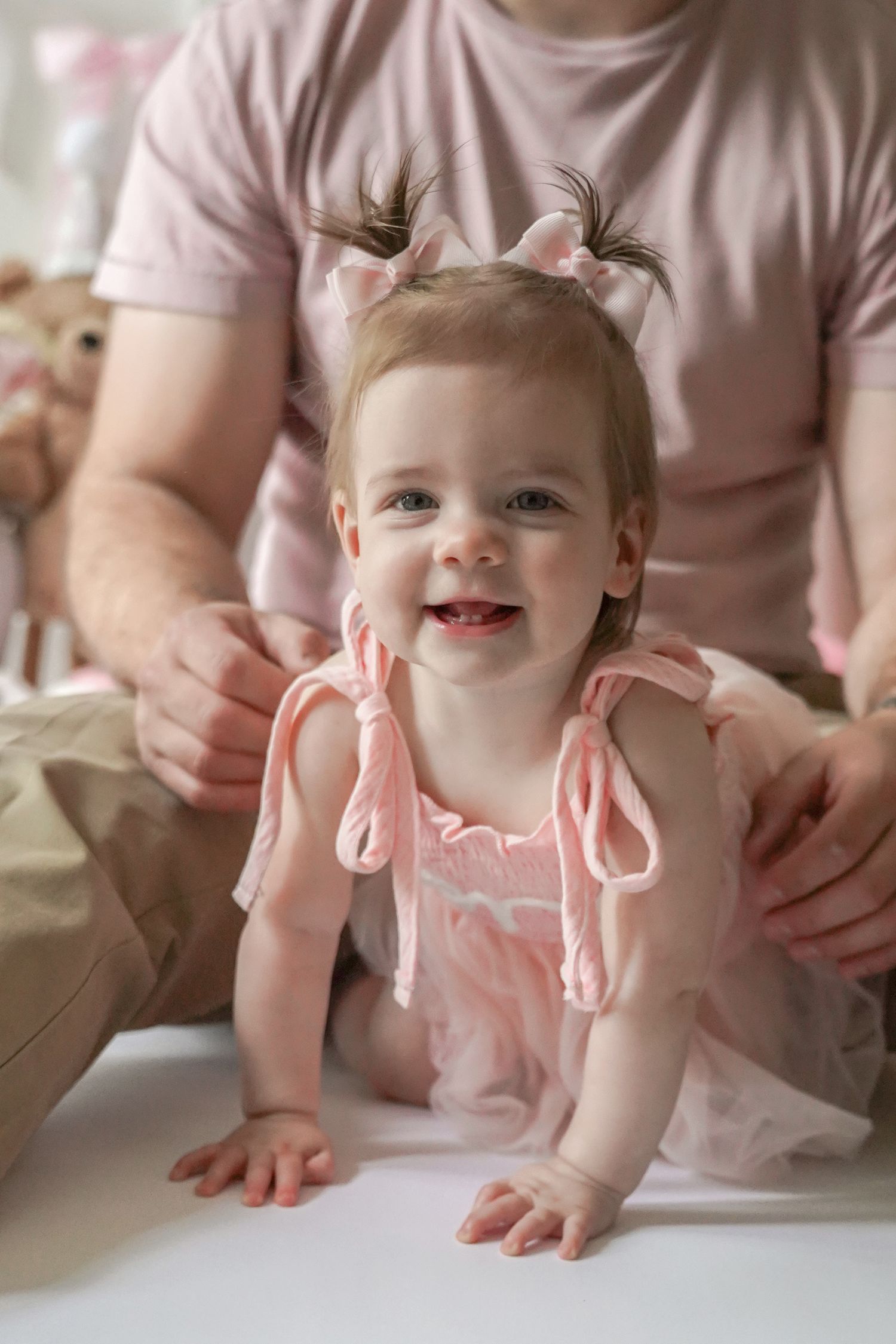 A happy baby wearing a pink dress with little pigtails sits on the floor with a caregiver in the background.