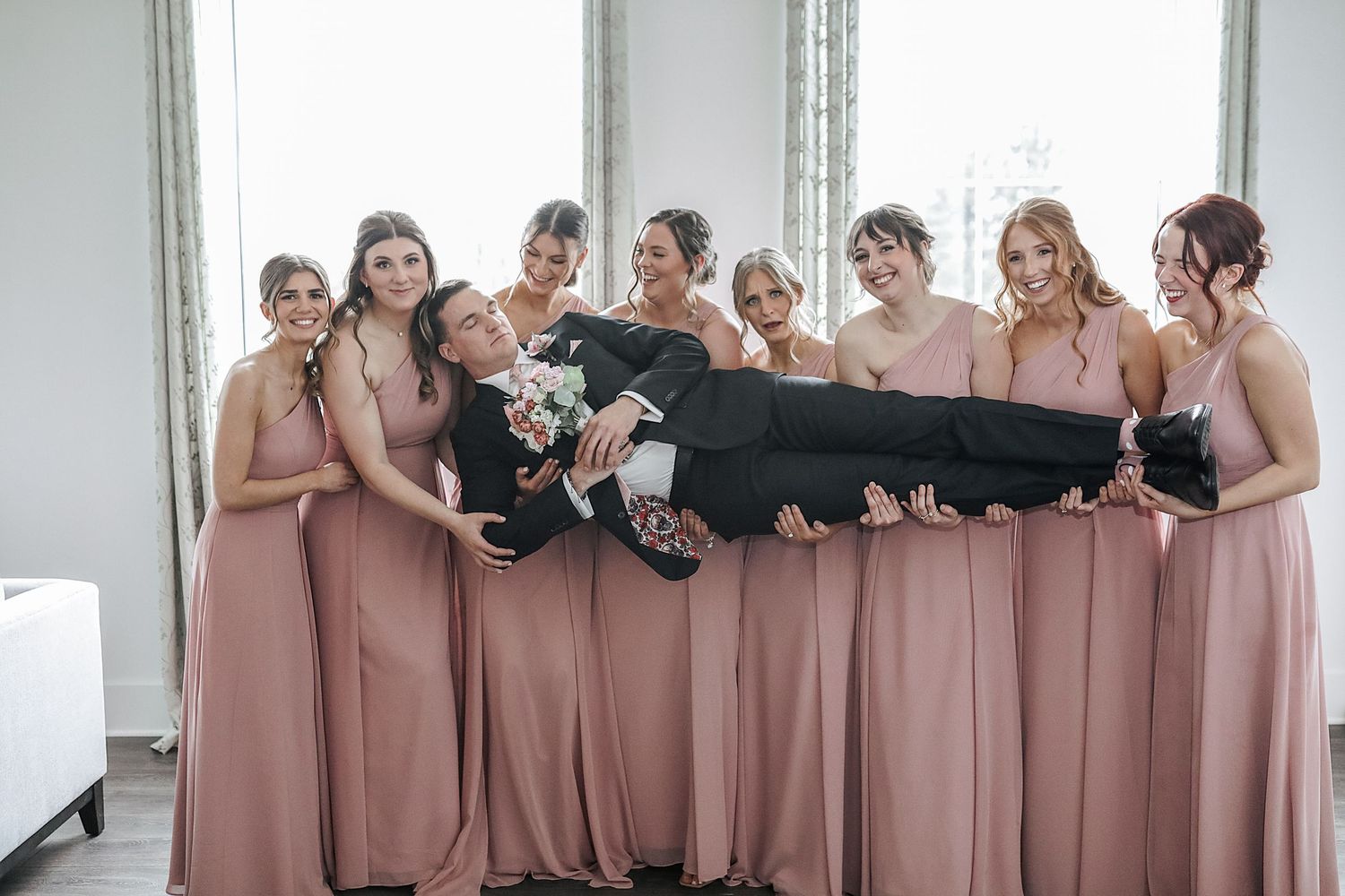 A group in matching dusty pink bridesmaid dresses hold someone wearing black horizontally while posing together.