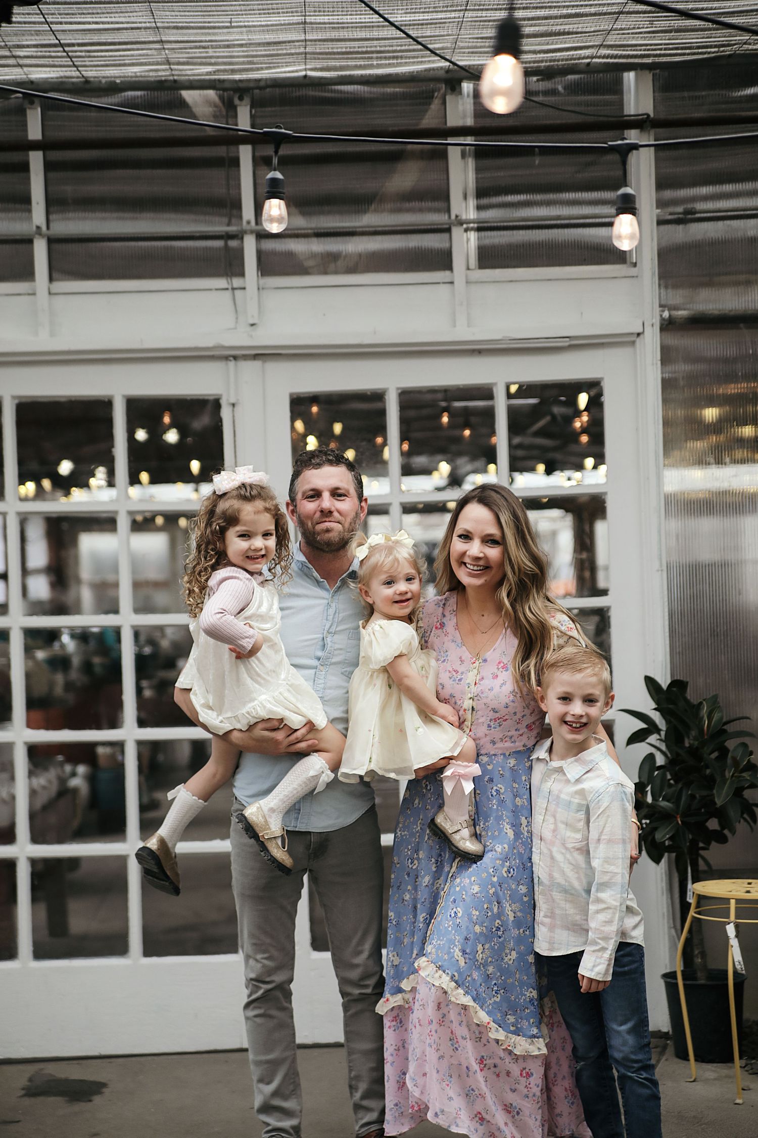 A family stands together in a bright indoor space with string lights and white walls.