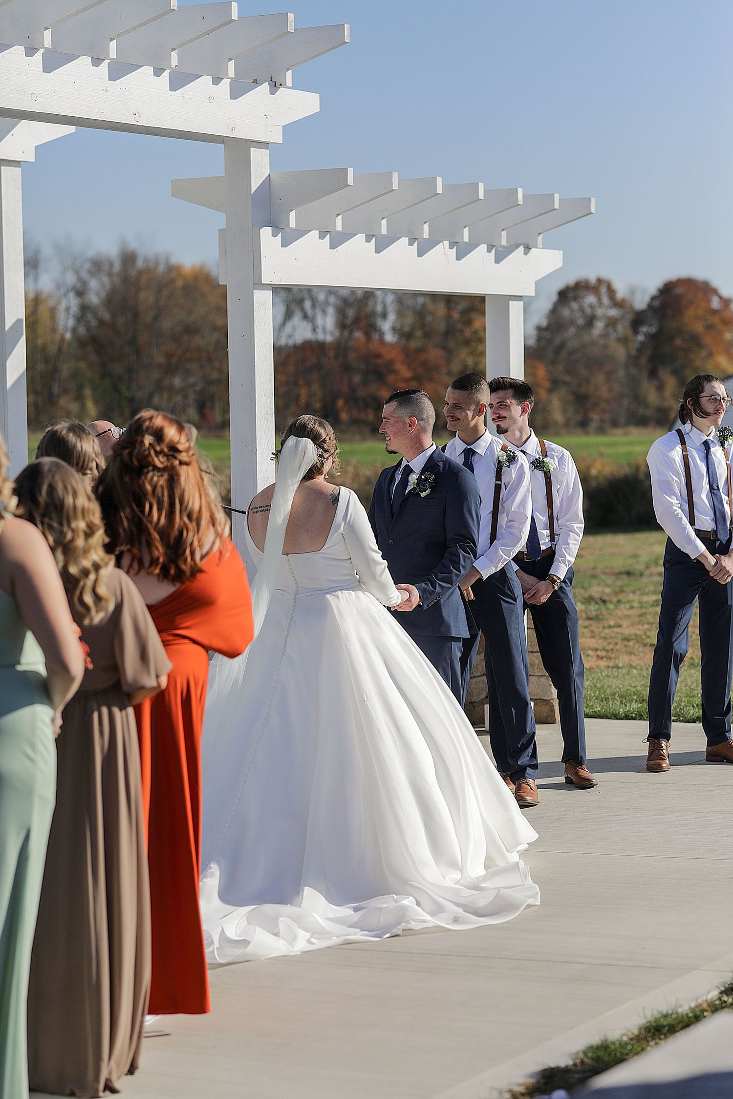 Outdoor wedding ceremony under white pergola with bridal party in colorful dresses and groomsmen in suspenders.