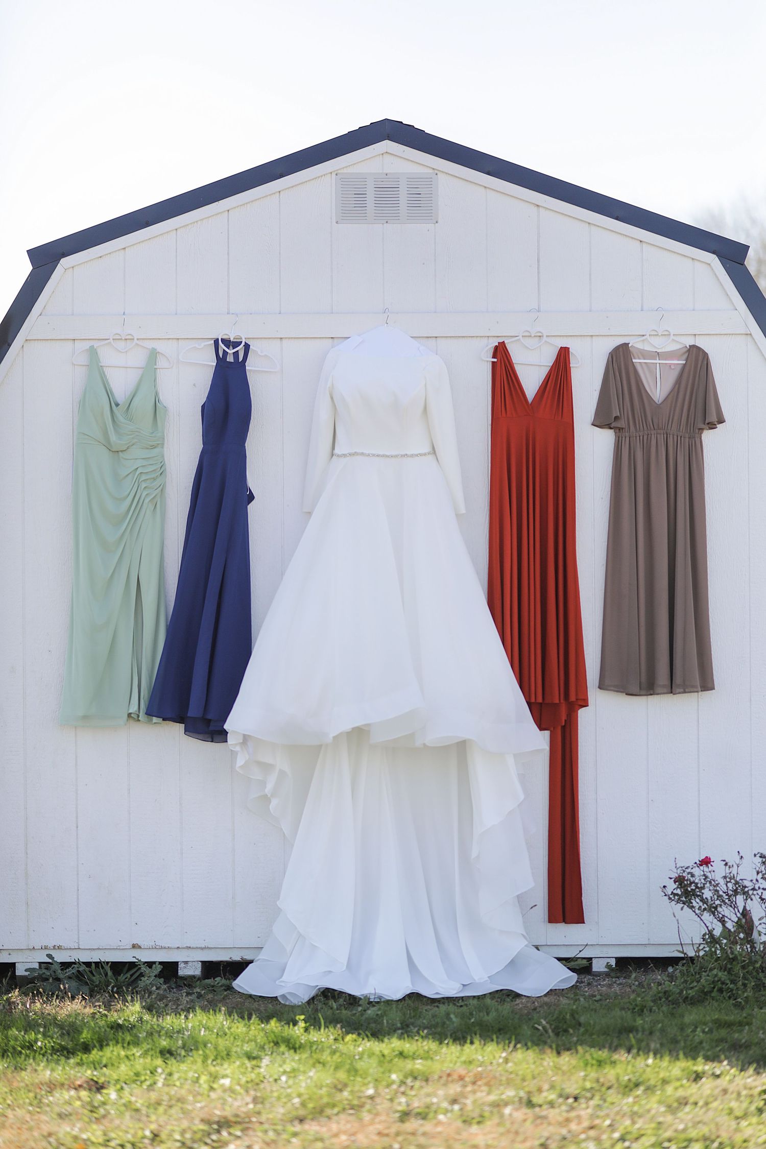 Wedding party dresses in mint green, navy blue, white, red and taupe hang on a white barn wall.