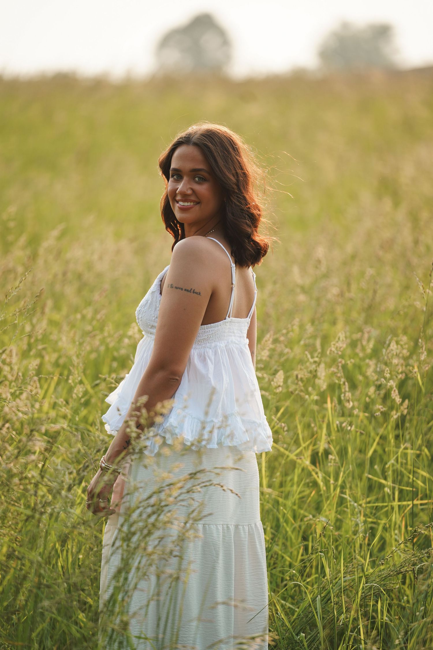 A figure in a flowing white summer dress poses gracefully in a sunlit meadow of tall grass.