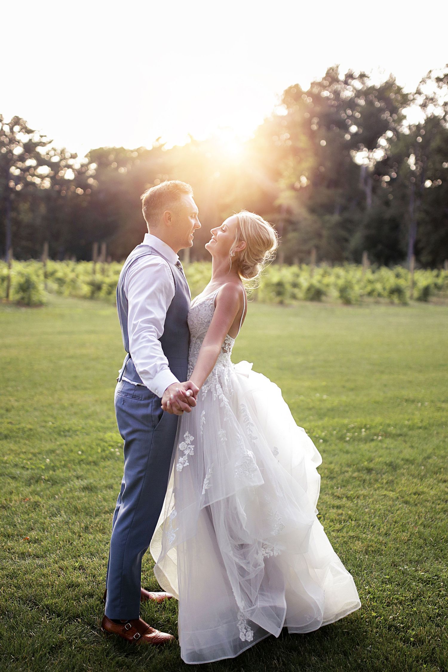 Romantic sunset portrait of newlyweds in an elegant white gown and blue vest on grassy field.