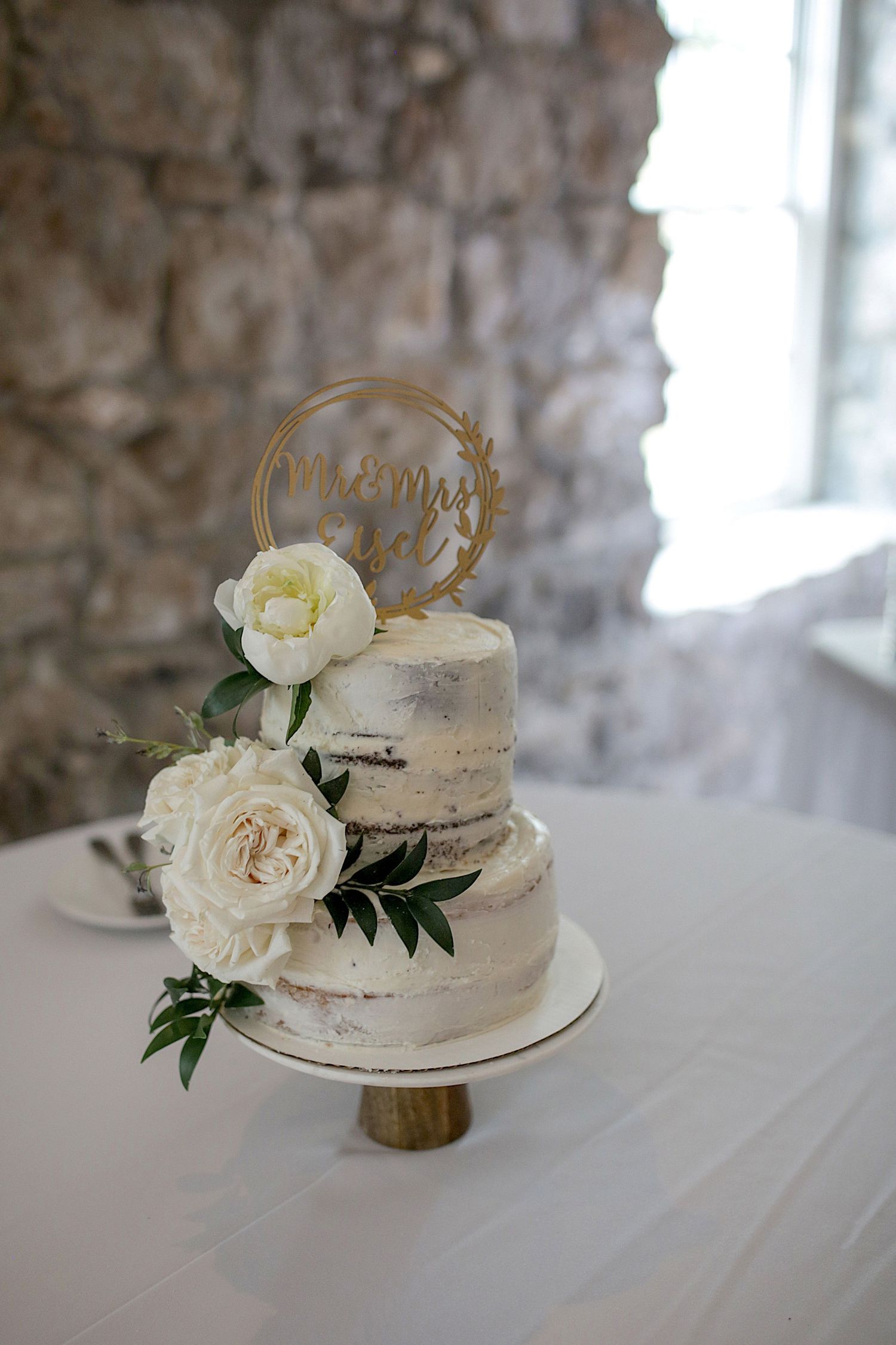 Rustic semi-naked wedding cake decorated with white roses and gold topper against stone wall.