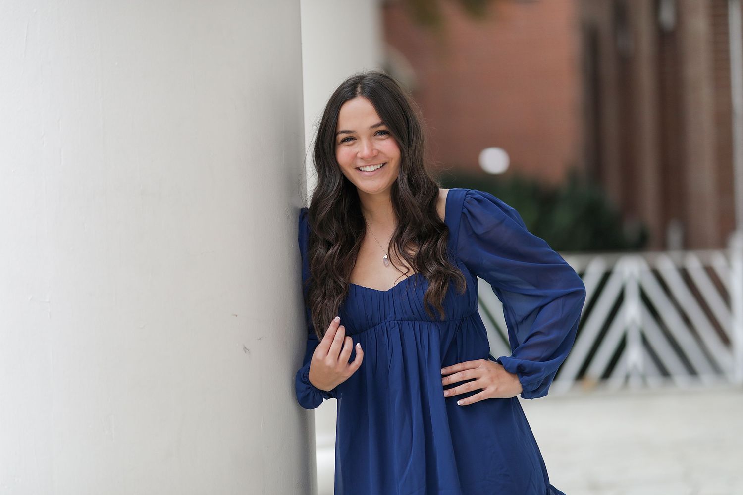 A woman in a navy blue long-sleeved dress poses against a white column outdoors.