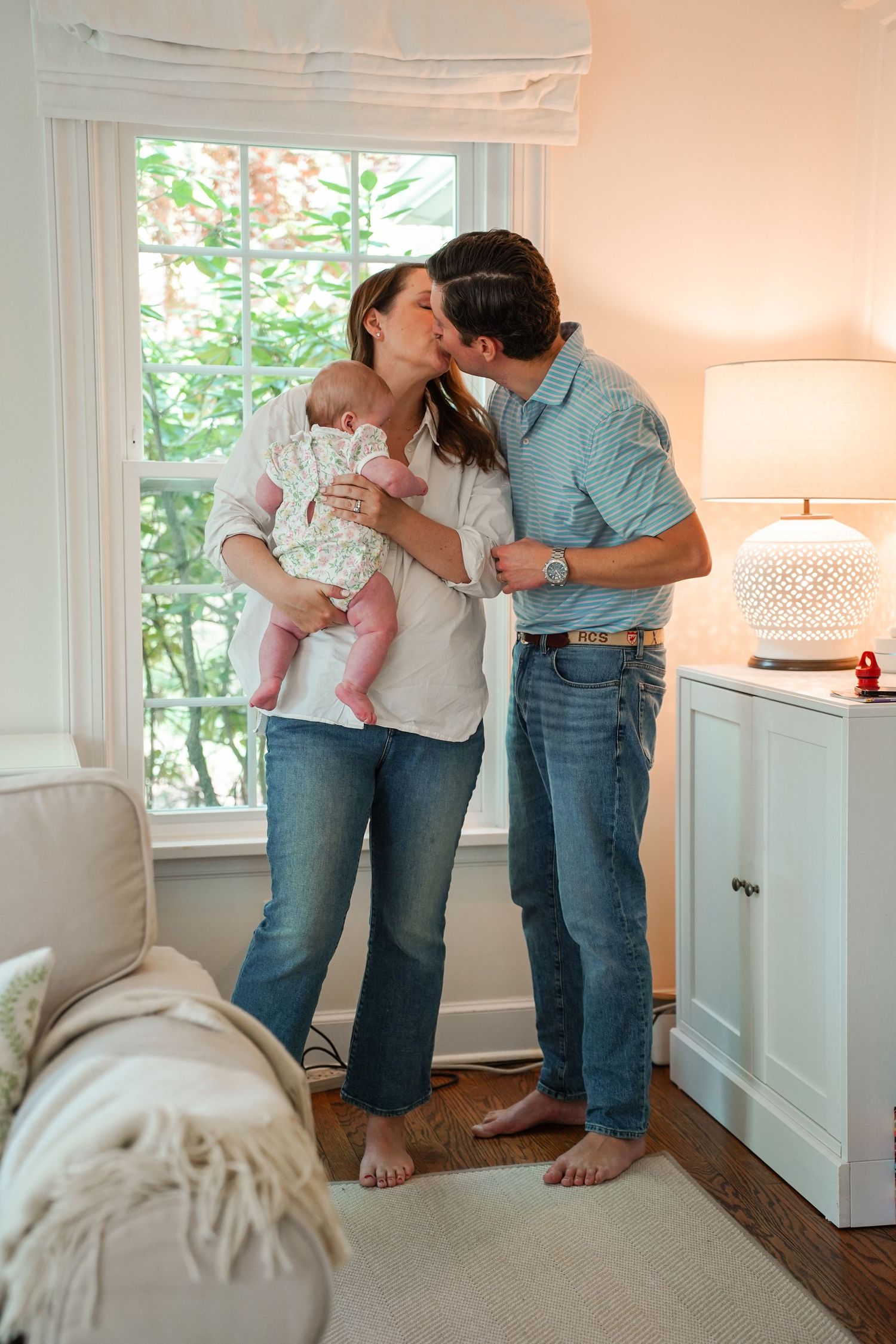 A couple shares an intimate moment while holding their baby in a sunlit corner of their home by a window with a white lamp.