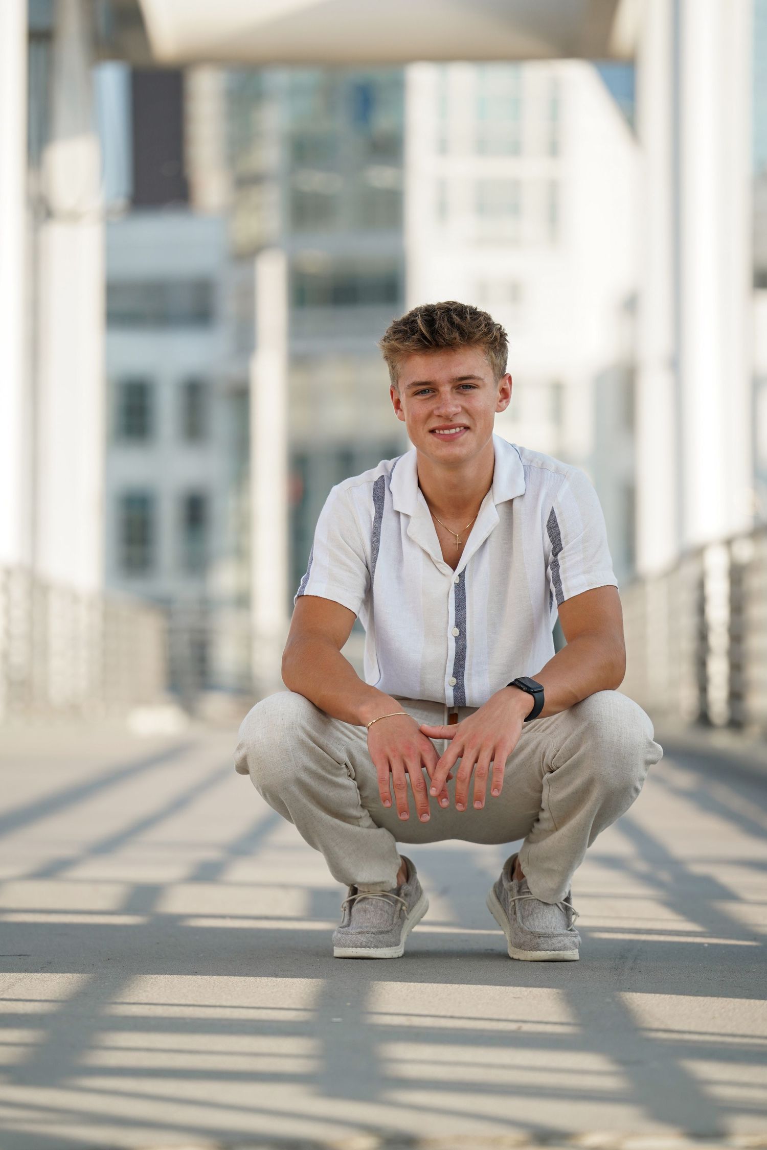 A person in a white shirt and khaki pants crouches on a sunlit walkway with modern architecture in the background.