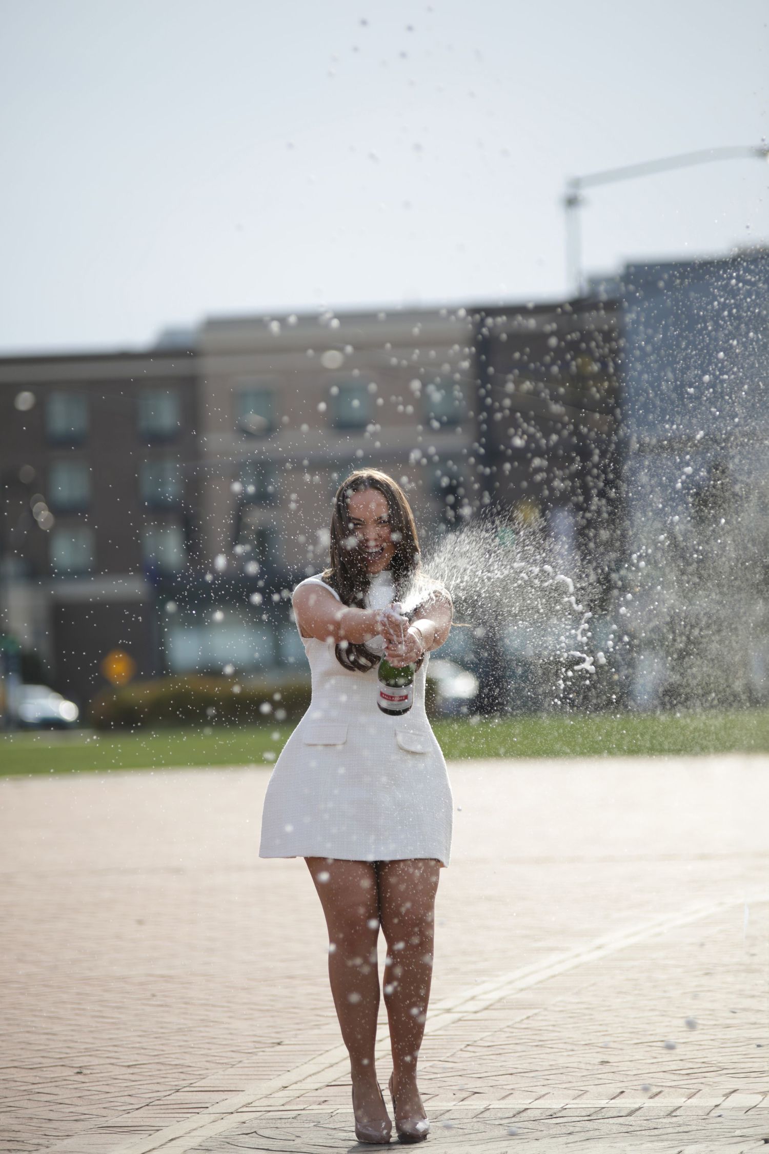 A person in a white dress stands on a paved area while water droplets splash around them in the sunlight.