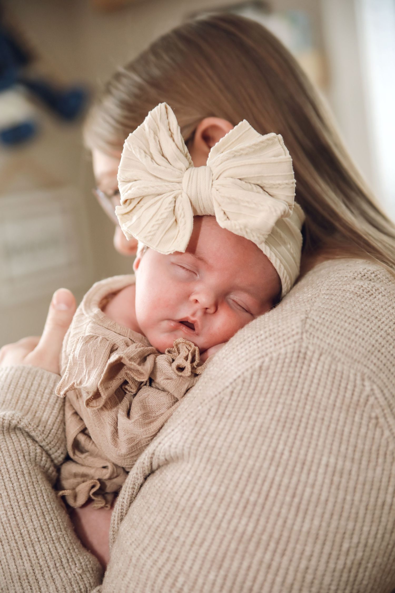 A sleeping newborn wearing a cream bow headband rests peacefully on a textured beige blanket in a series of photos.