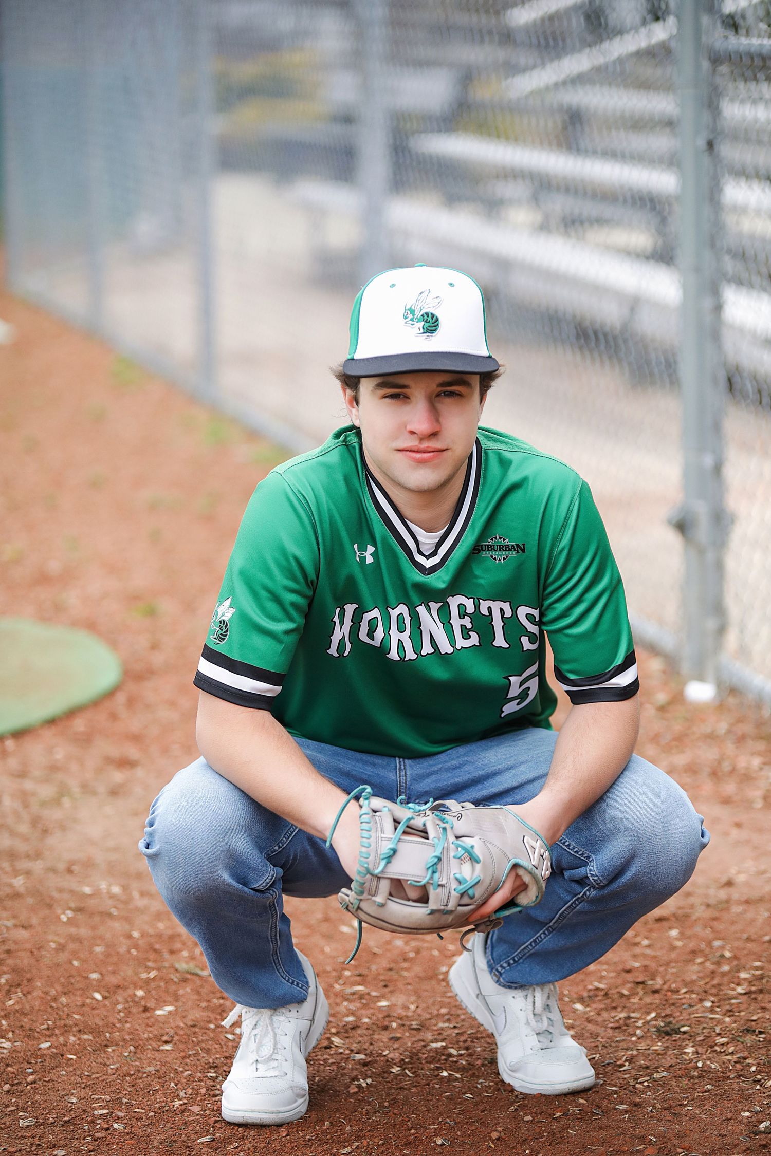 Baseball player in green Hornets jersey and white cap crouches in dugout during game.