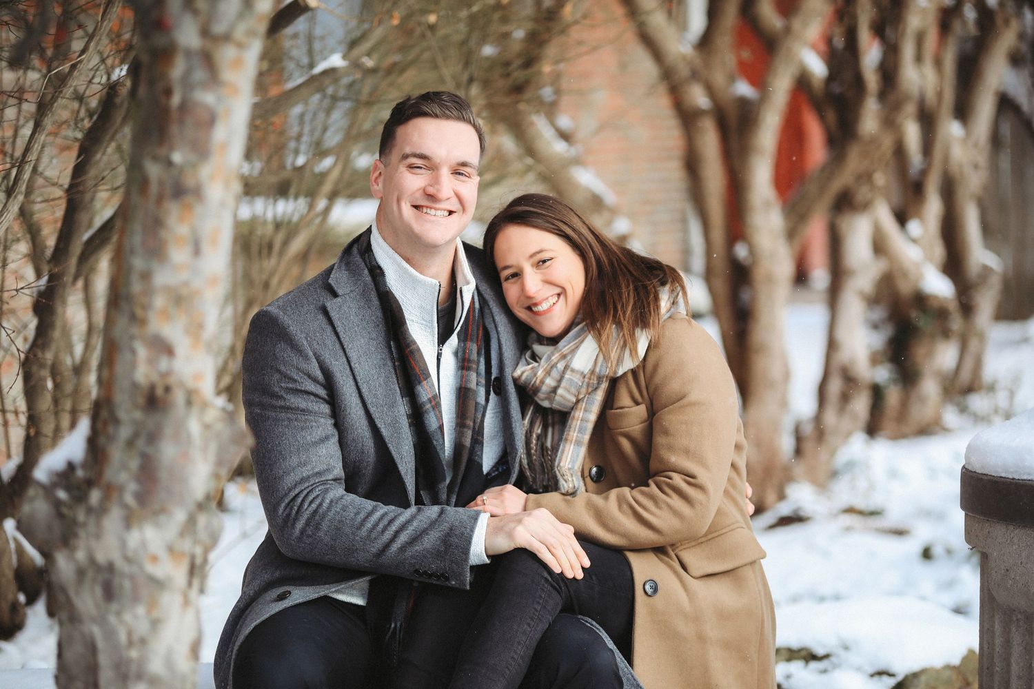 A couple sits together on a snowy winter day in a park setting, wearing warm coats and sharing intimate moments.