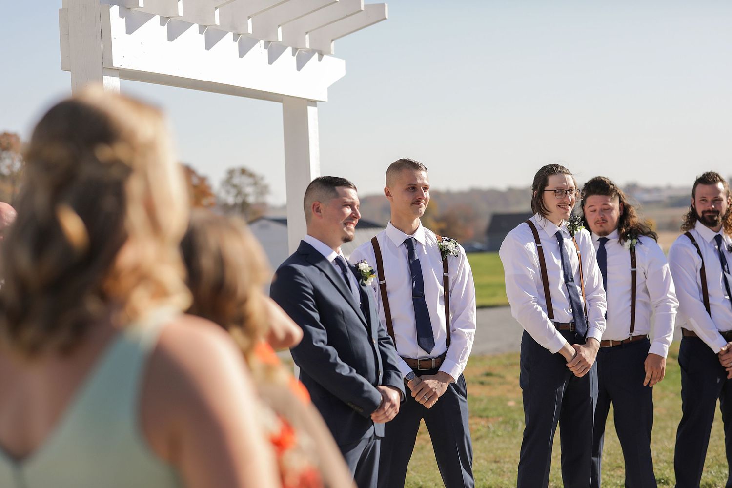 Wedding party stands under white pergola during outdoor ceremony on sunny autumn day.