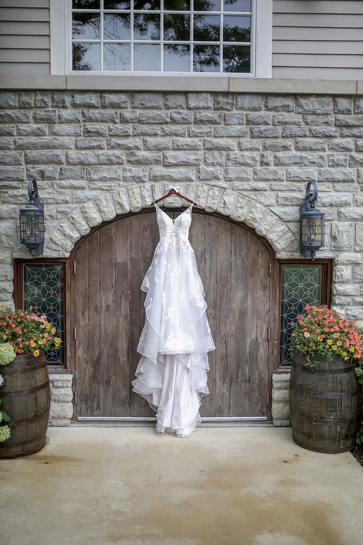 White wedding dress hanging against rustic wooden doors and stone wall with flowers in barrels.