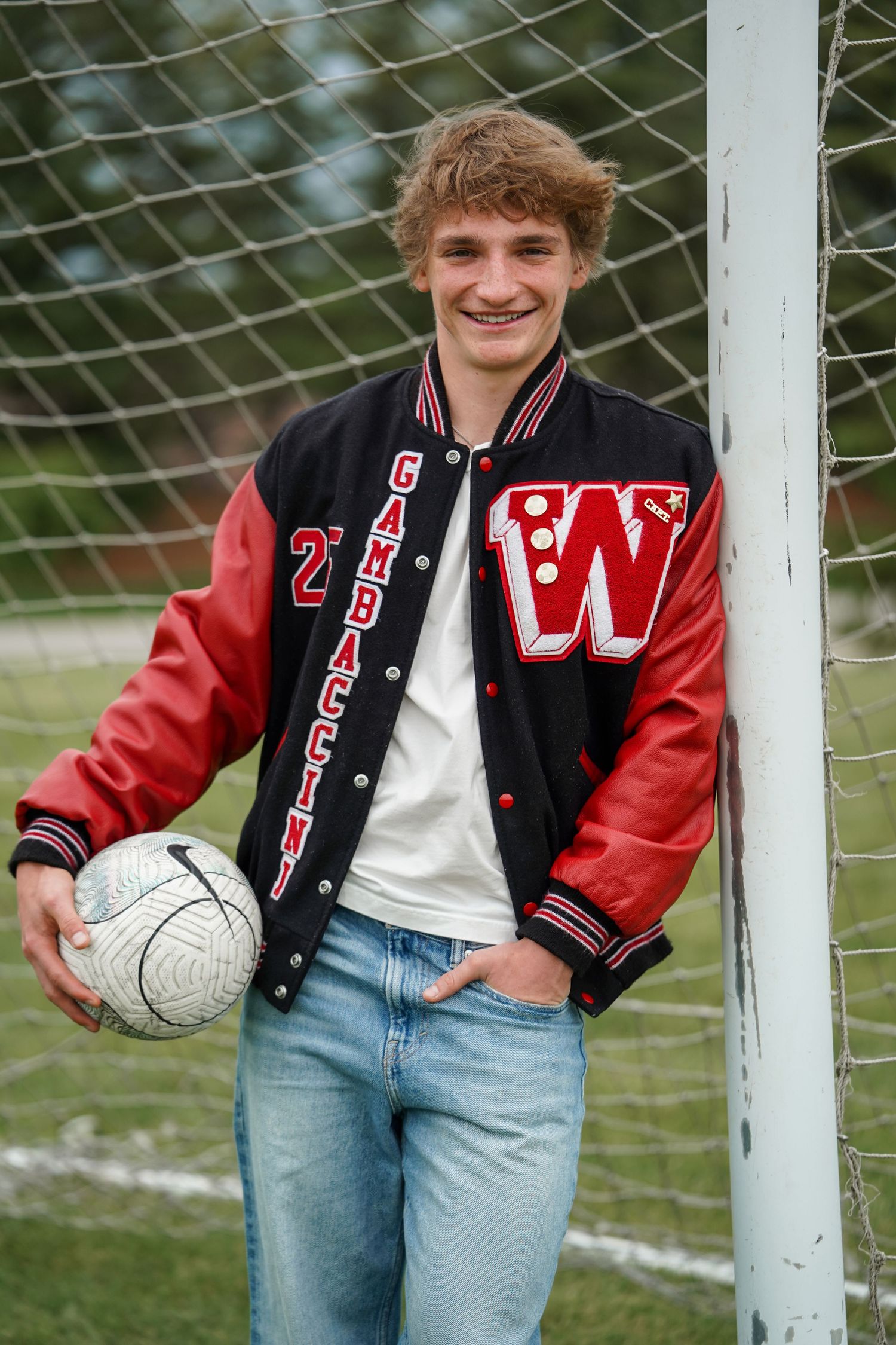 High school athlete in varsity letterman jacket poses with a soccer ball by goal post on sports field.