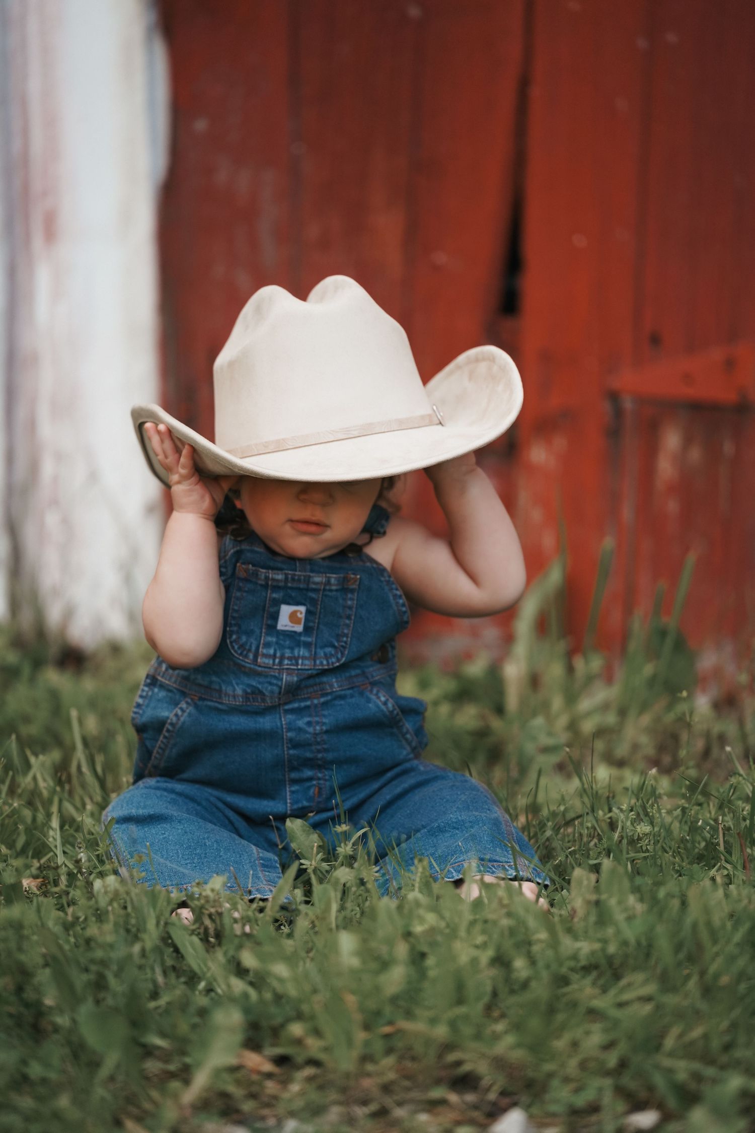 A small child in denim overalls sits in grass wearing an oversized white cowboy hat near a red barn wall.