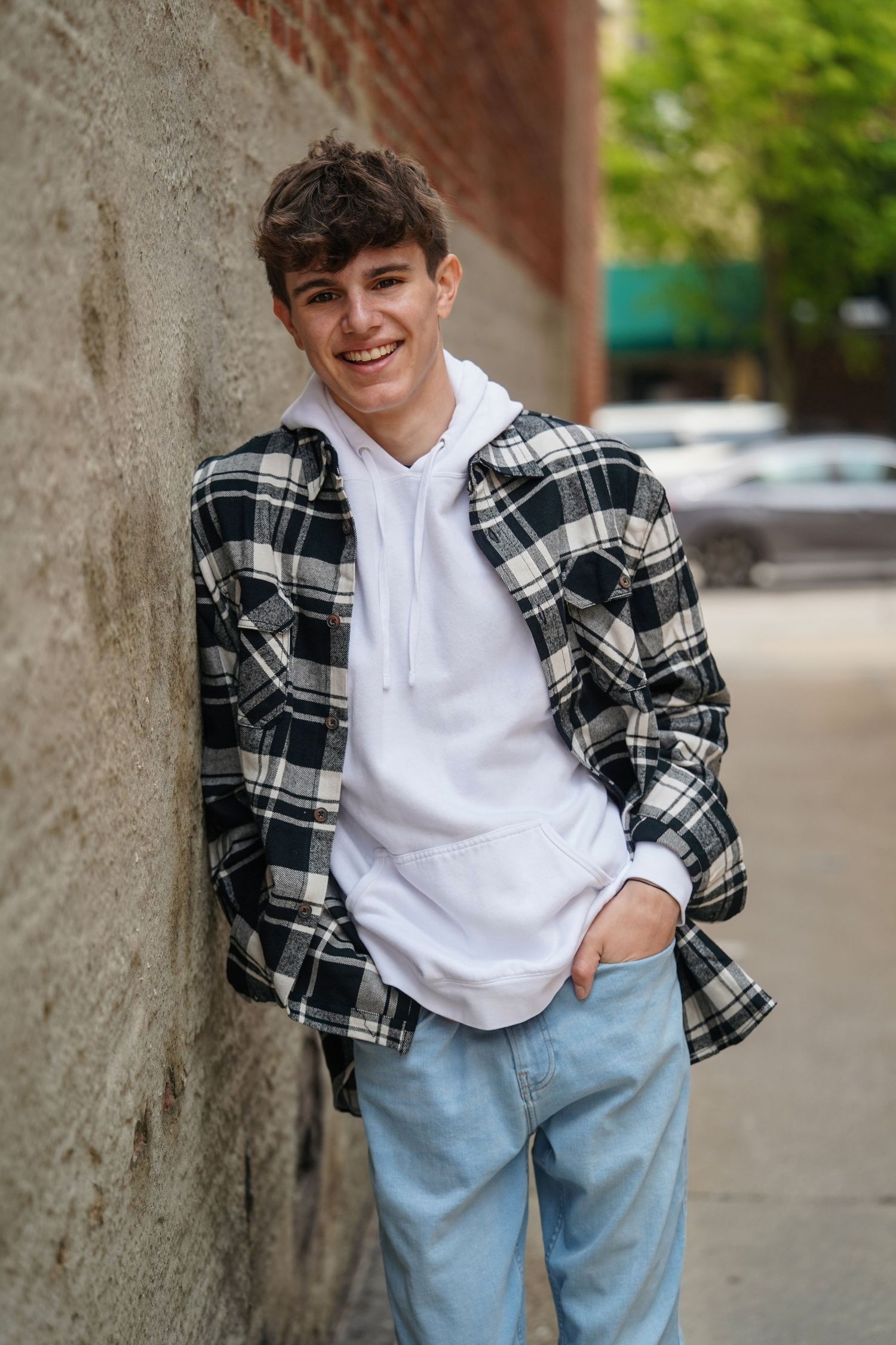 Casual street style outfit featuring black plaid flannel shirt over white hoodie with light wash jeans against brick wall.