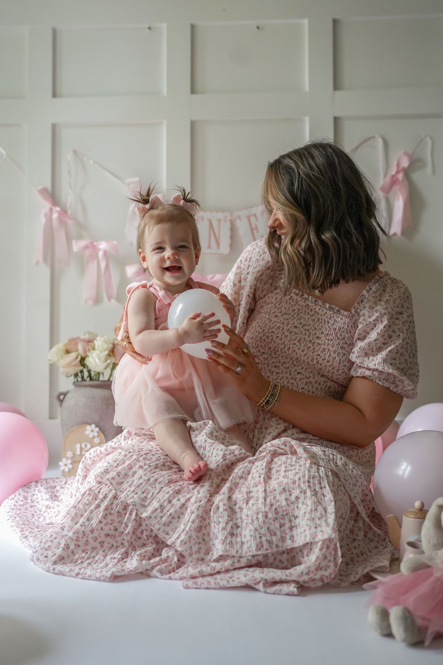 Parent and baby in matching pink floral dresses share joyful moments during a pink-themed photo session with balloons.
