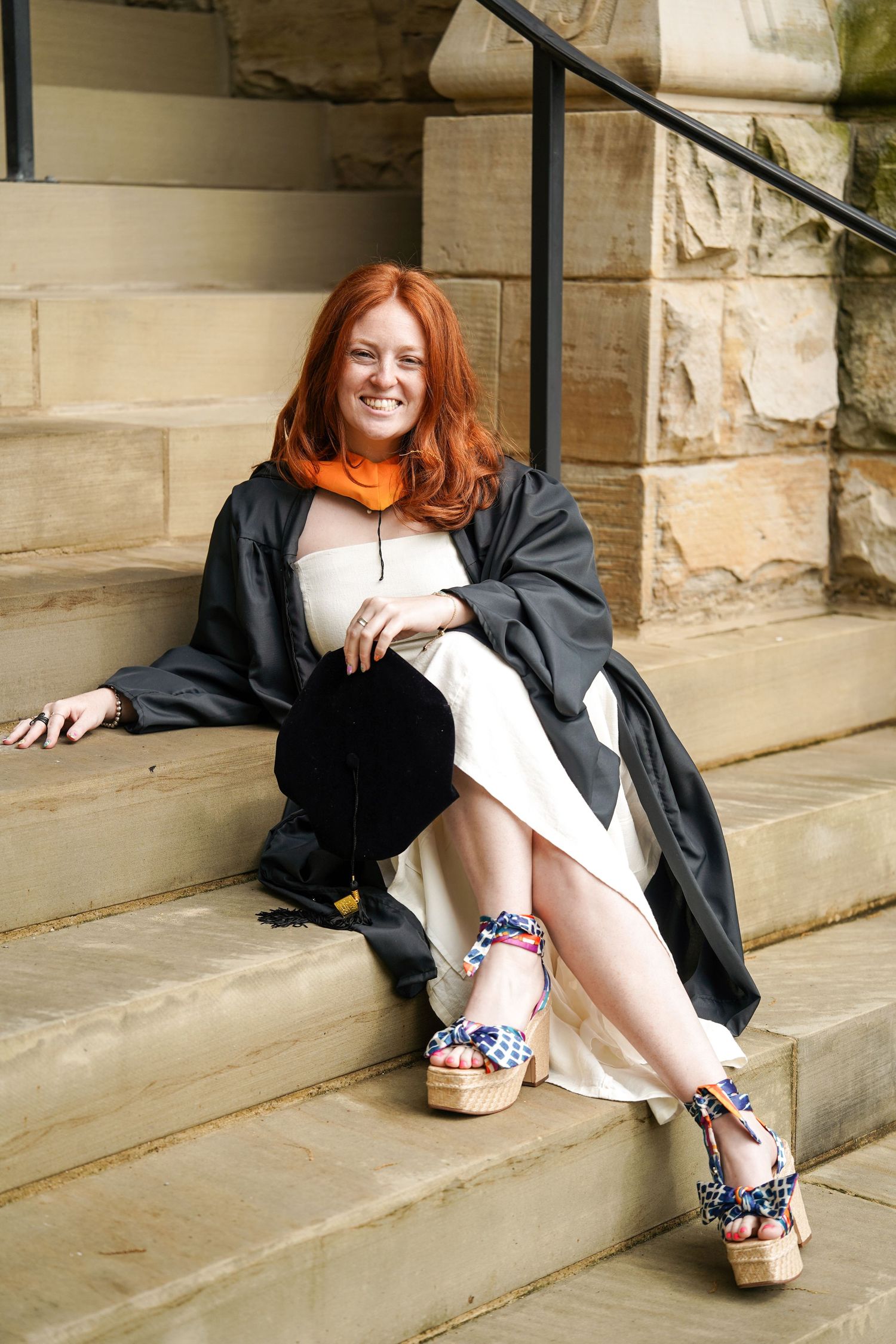 A person in a black graduation gown sits casually on concrete steps against a stone wall background.