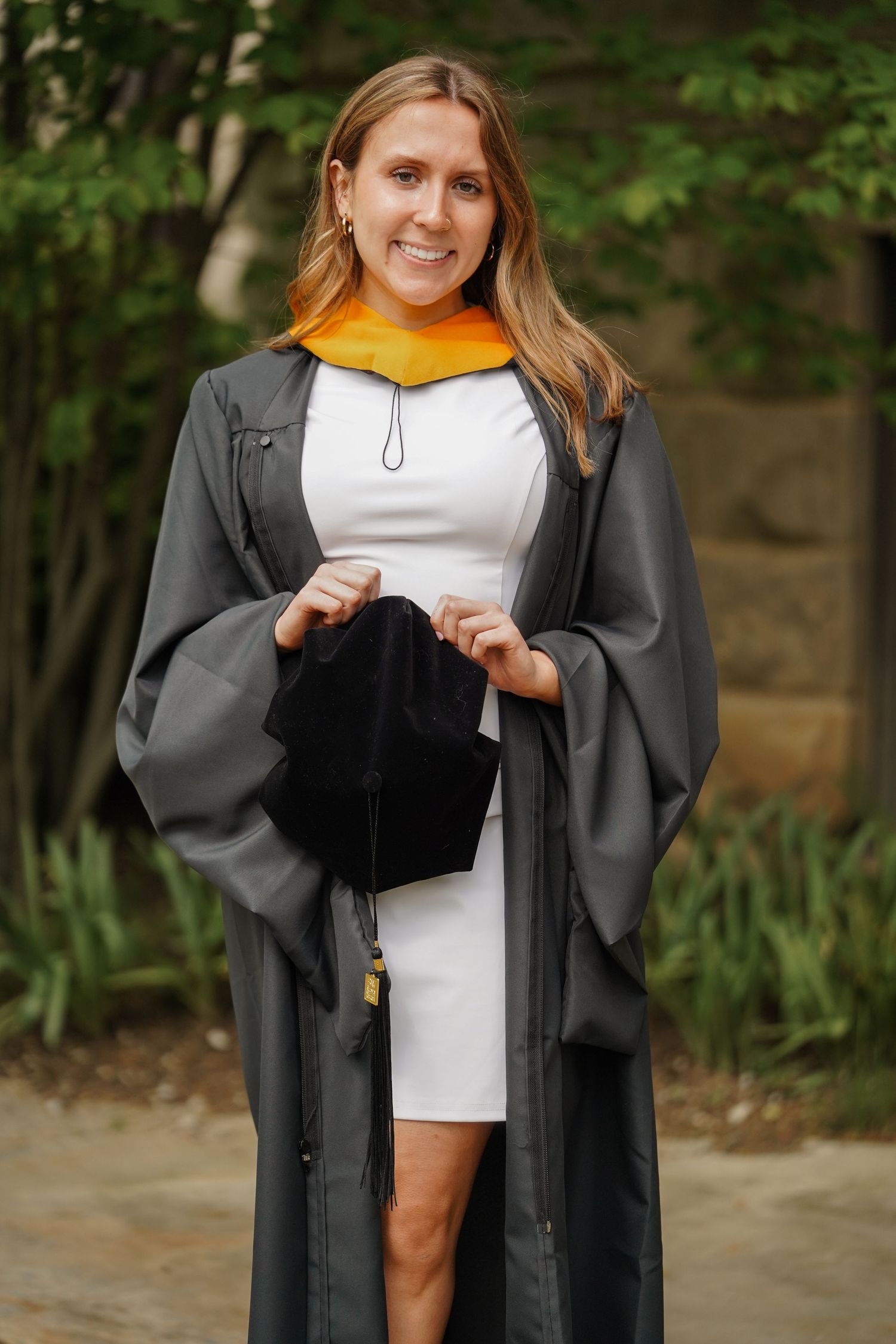 Graduate in cap and gown holds black graduation cap during outdoor graduation ceremony.
