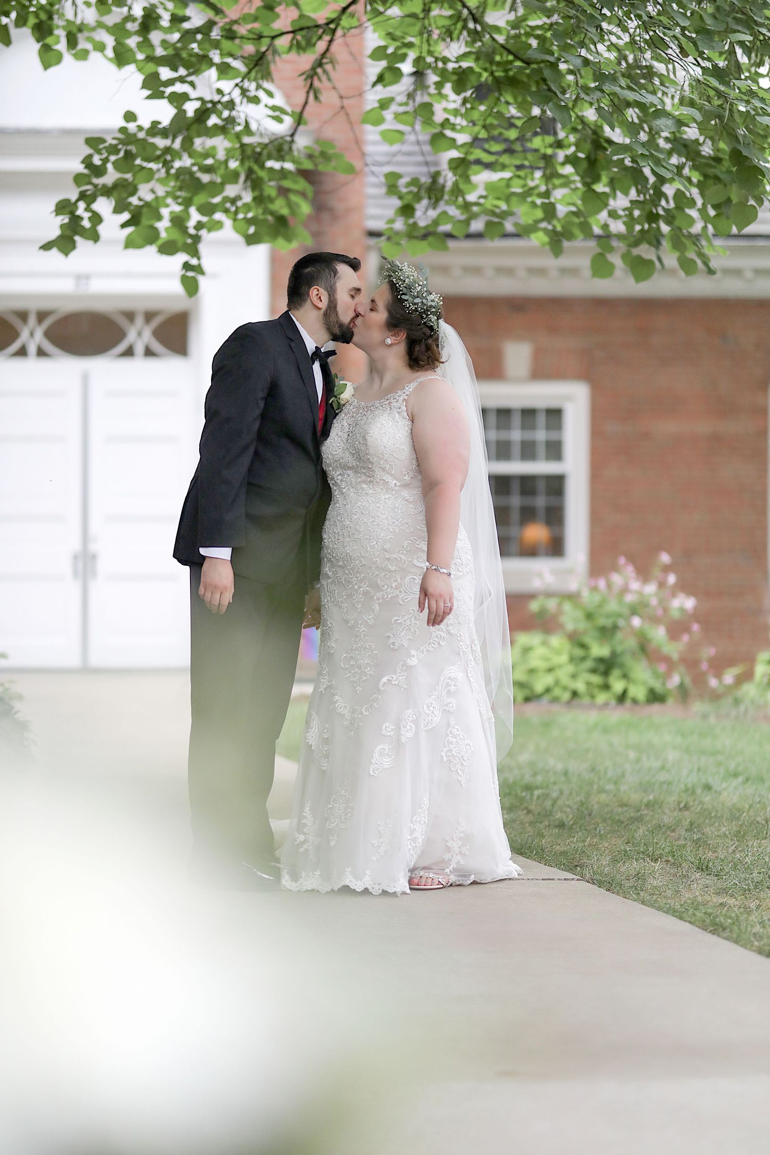 A romantic wedding moment under green trees outside a red brick building with white columns and architectural details.
