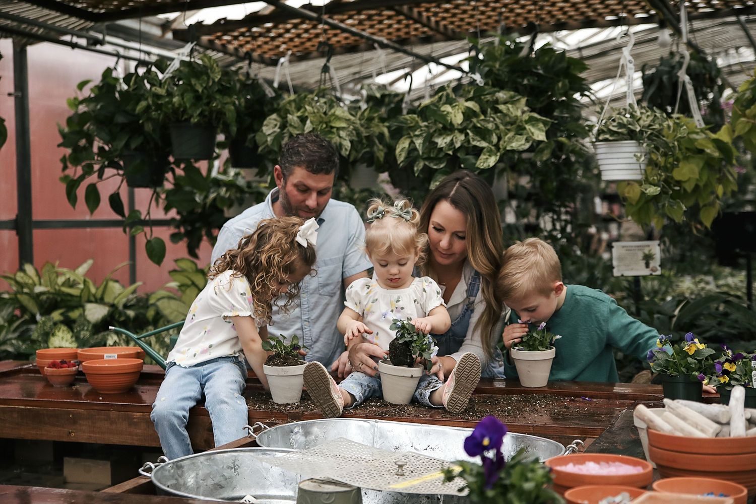 Family spends time together gardening at a greenhouse table with clay pots and flowers.