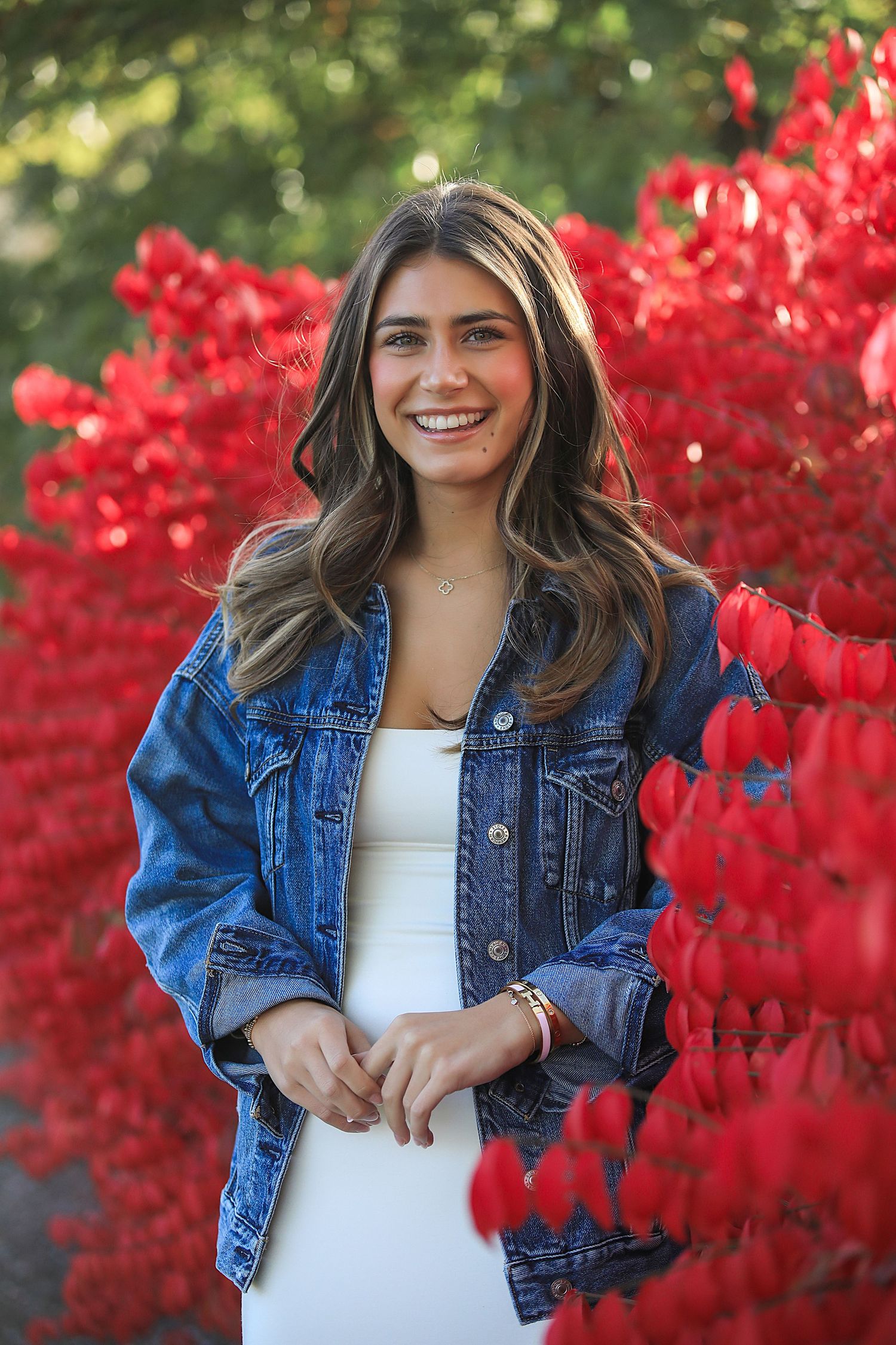 A portrait taken outdoors with bright red autumn foliage in the background, wearing a denim jacket and white top.