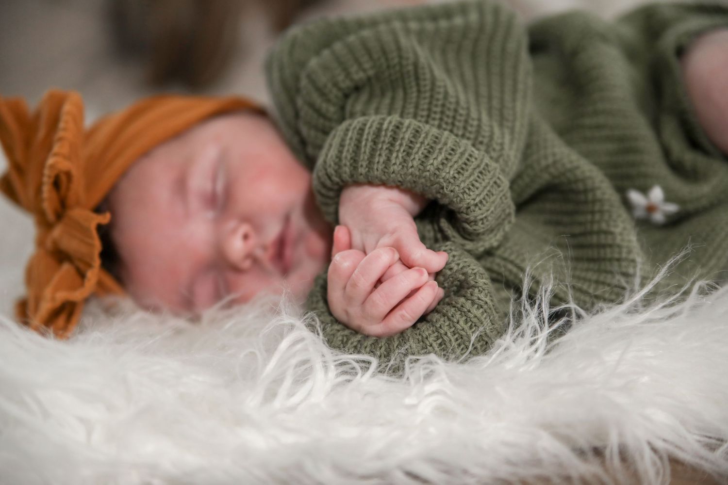 A baby wearing a green knit sweater lies on a soft white fur blanket while sleeping peacefully.