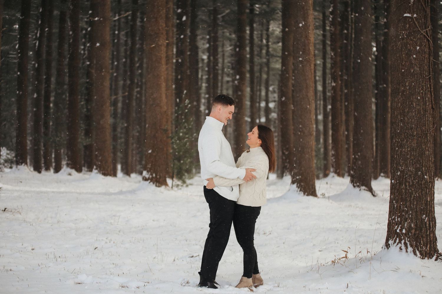 A couple shares a romantic moment in a snowy forest setting with tall pine trees in the background.