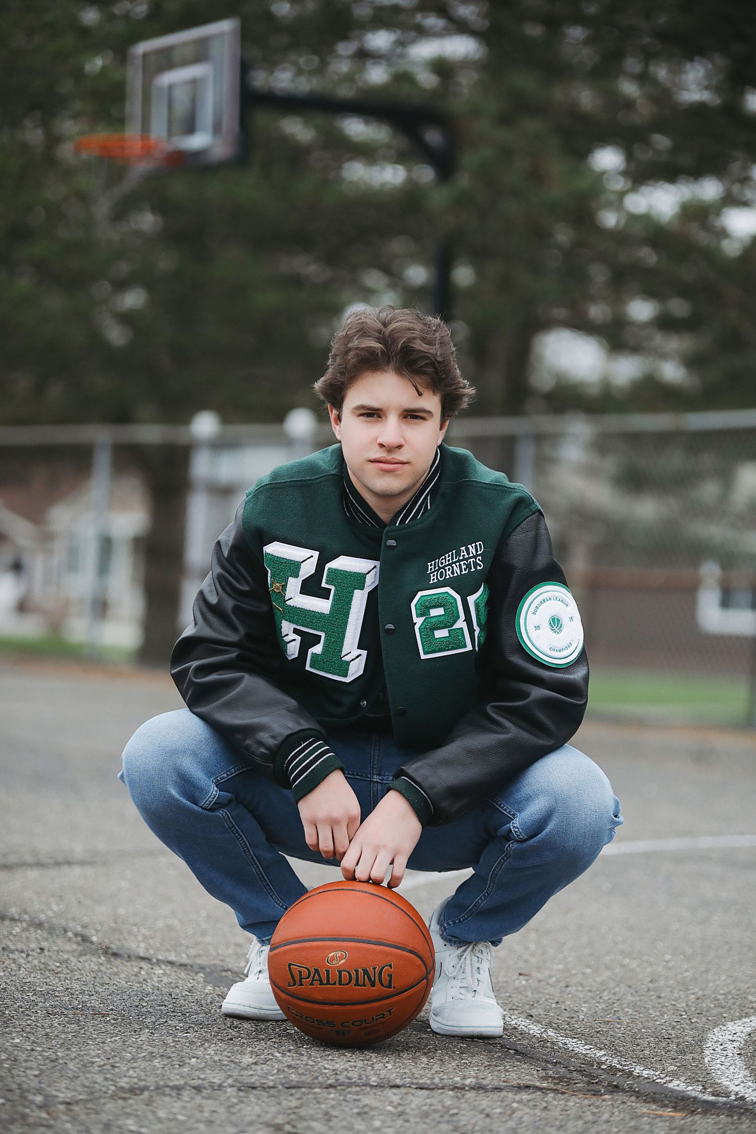 A basketball player in a green varsity jacket crouches on an outdoor court with a Spalding ball and hoop in background.