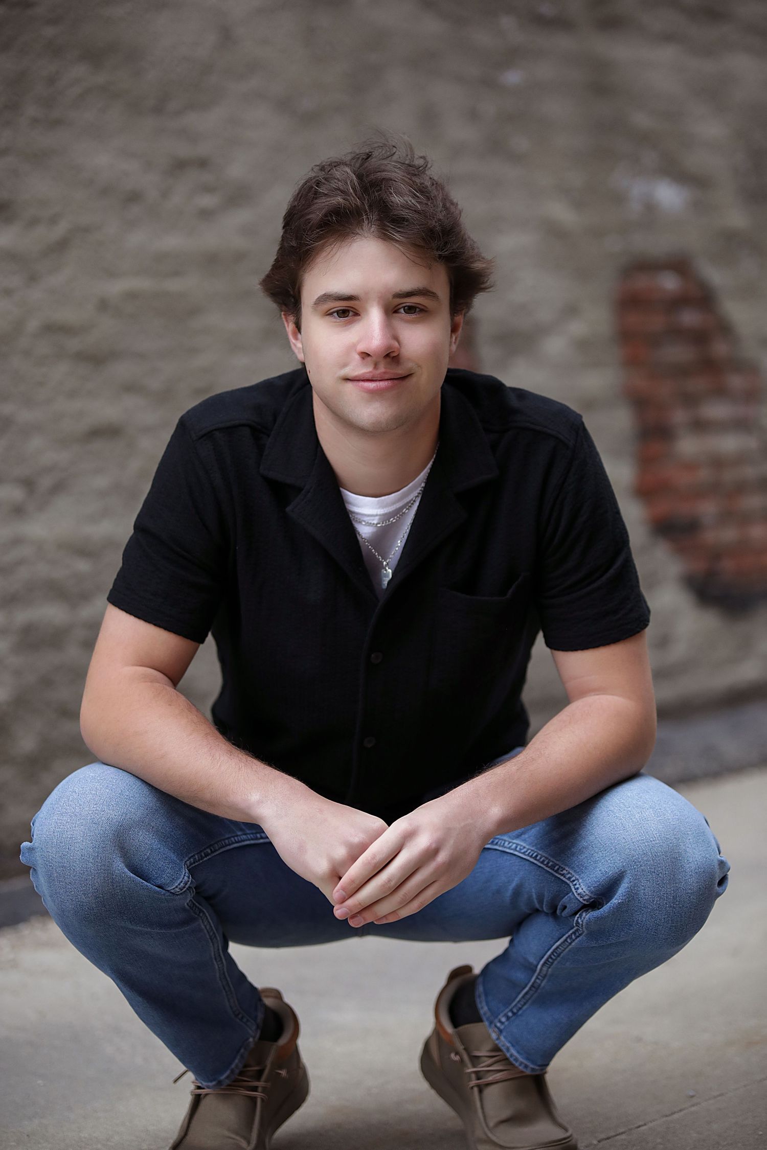 A young person in a black button-up shirt and jeans crouches casually against a concrete wall.