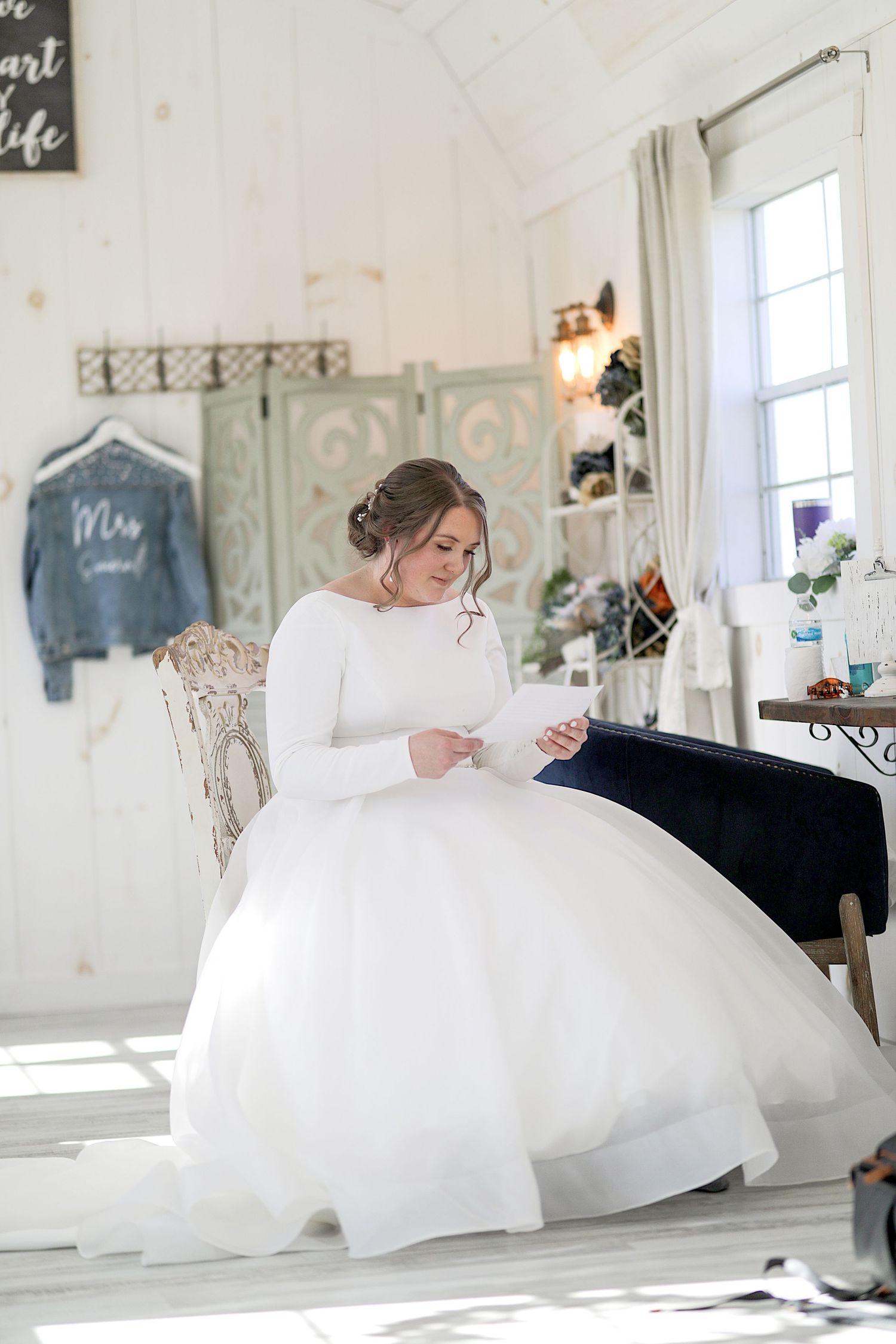 Bride in white long-sleeve gown reading in a rustic white room with vintage decor.