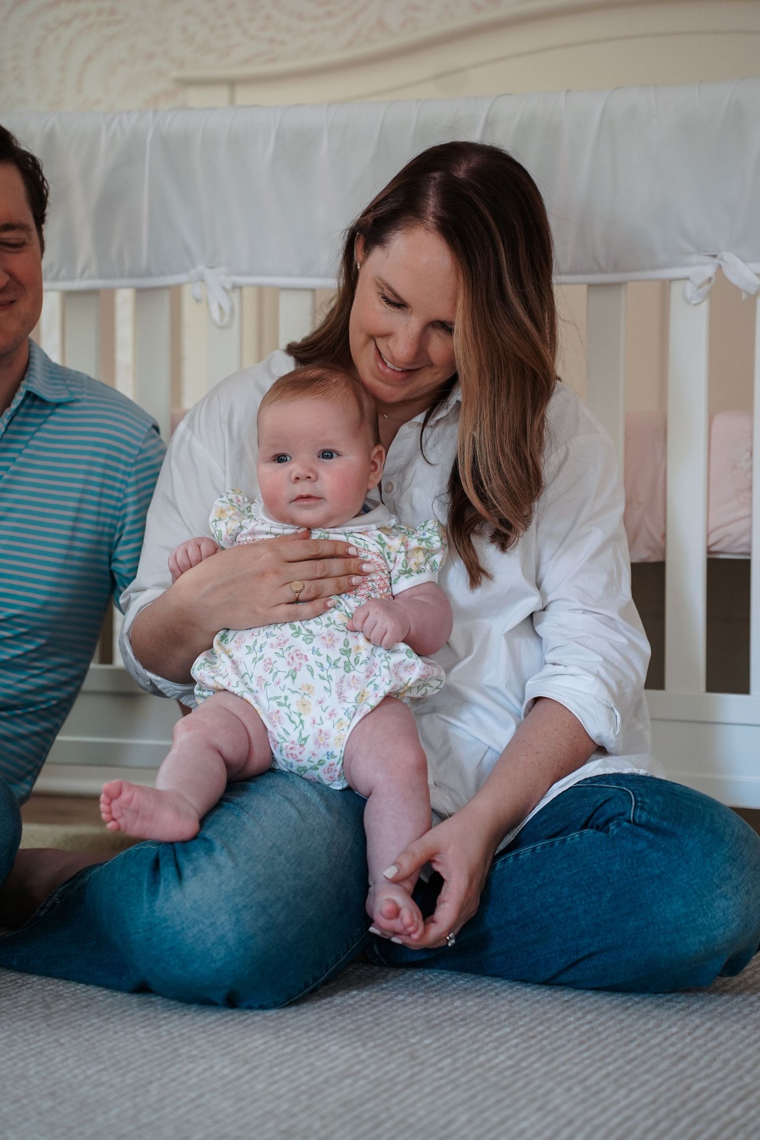 Baby in floral outfit lies on gray carpet while parent in white top and jeans sits nearby in modern nursery setting.