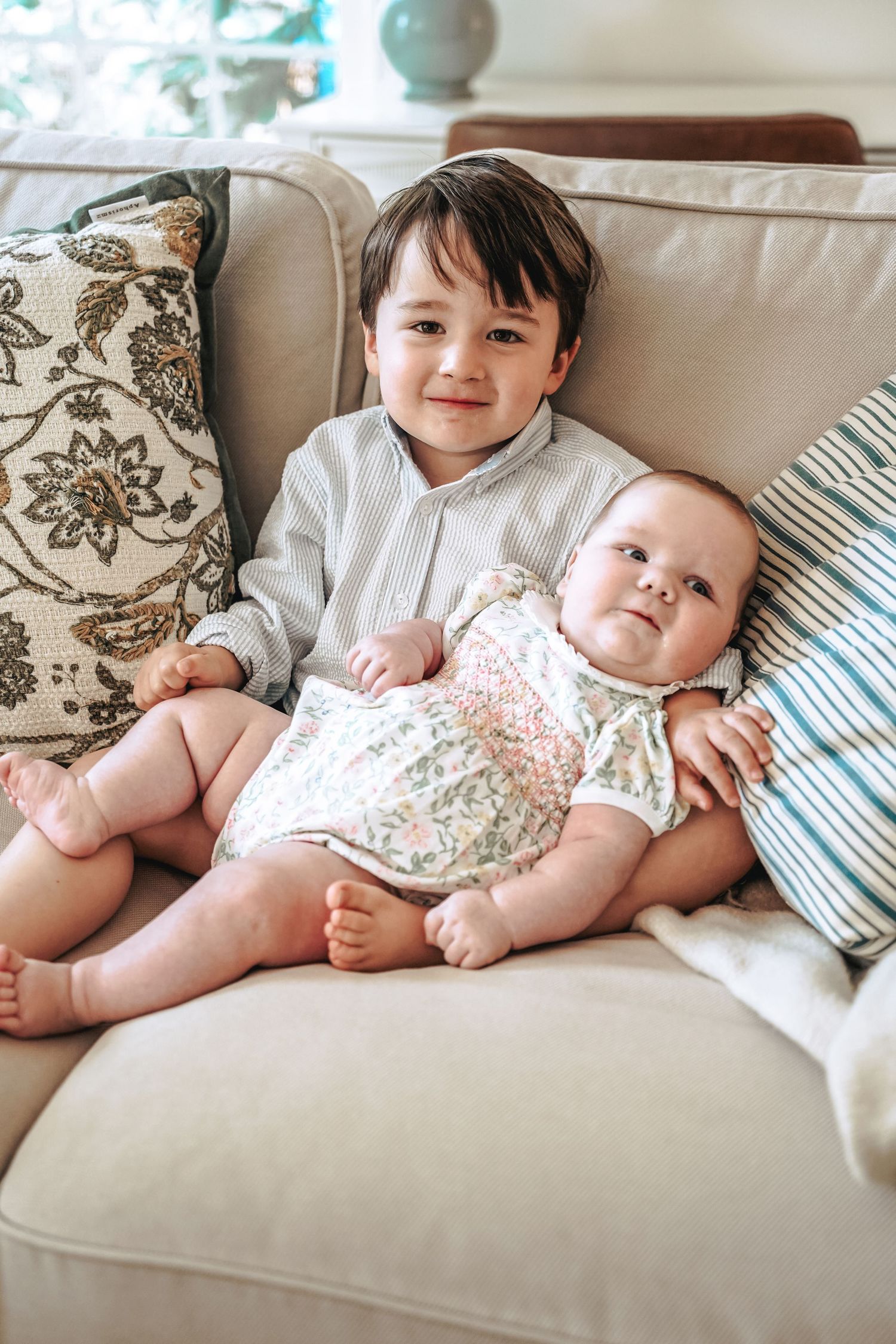 Two children relax together on a comfortable beige couch with patterned pillows in natural lighting.