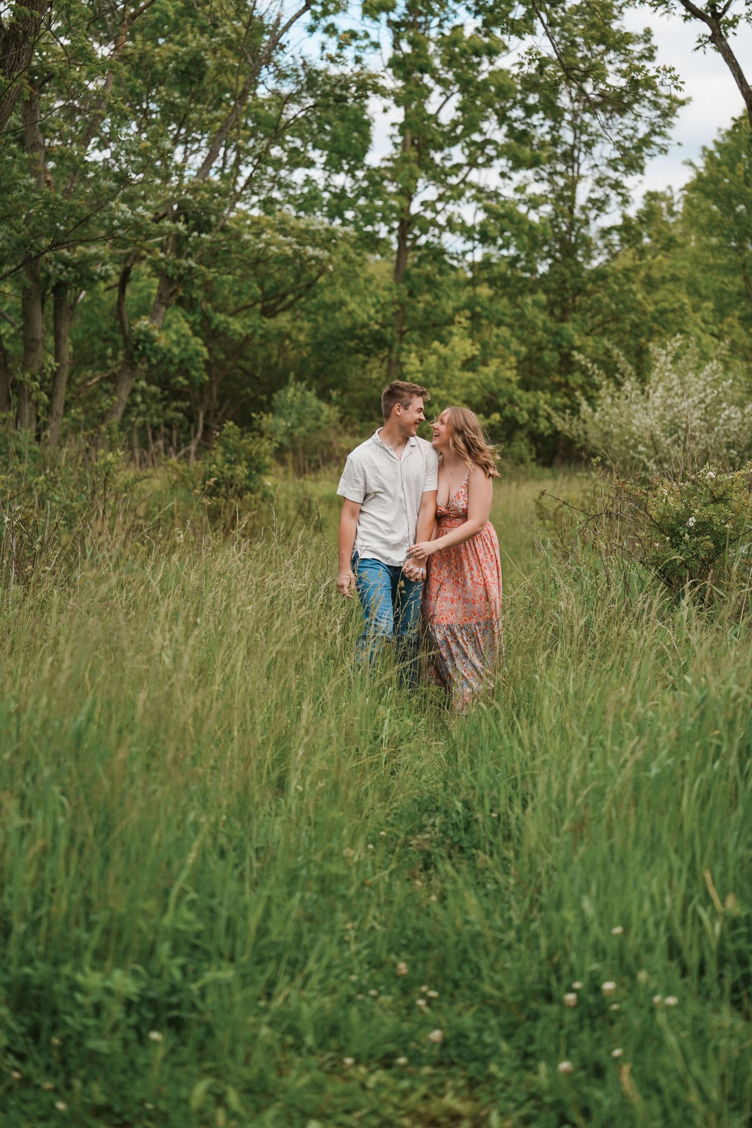 A couple walks intimately through tall summer grass and wildflowers in a romantic outdoor setting.