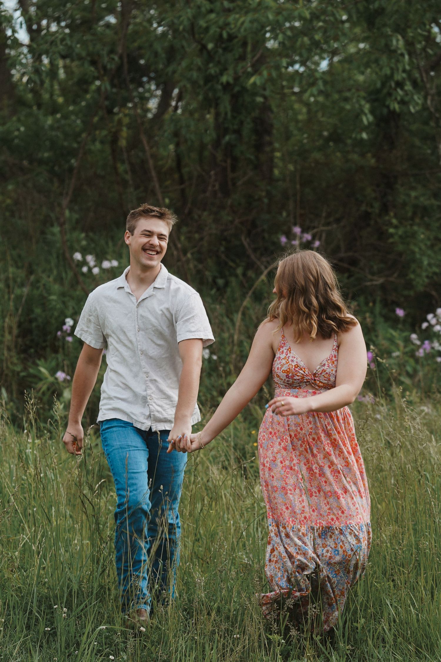 A couple walking hand in hand through a grassy field during a casual summer photoshoot.