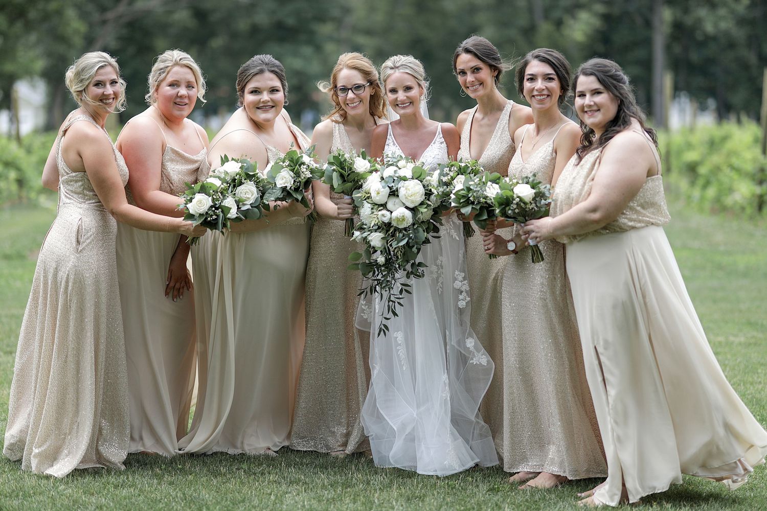 Bridesmaids in beige dresses posing together outdoors with floral bouquets.