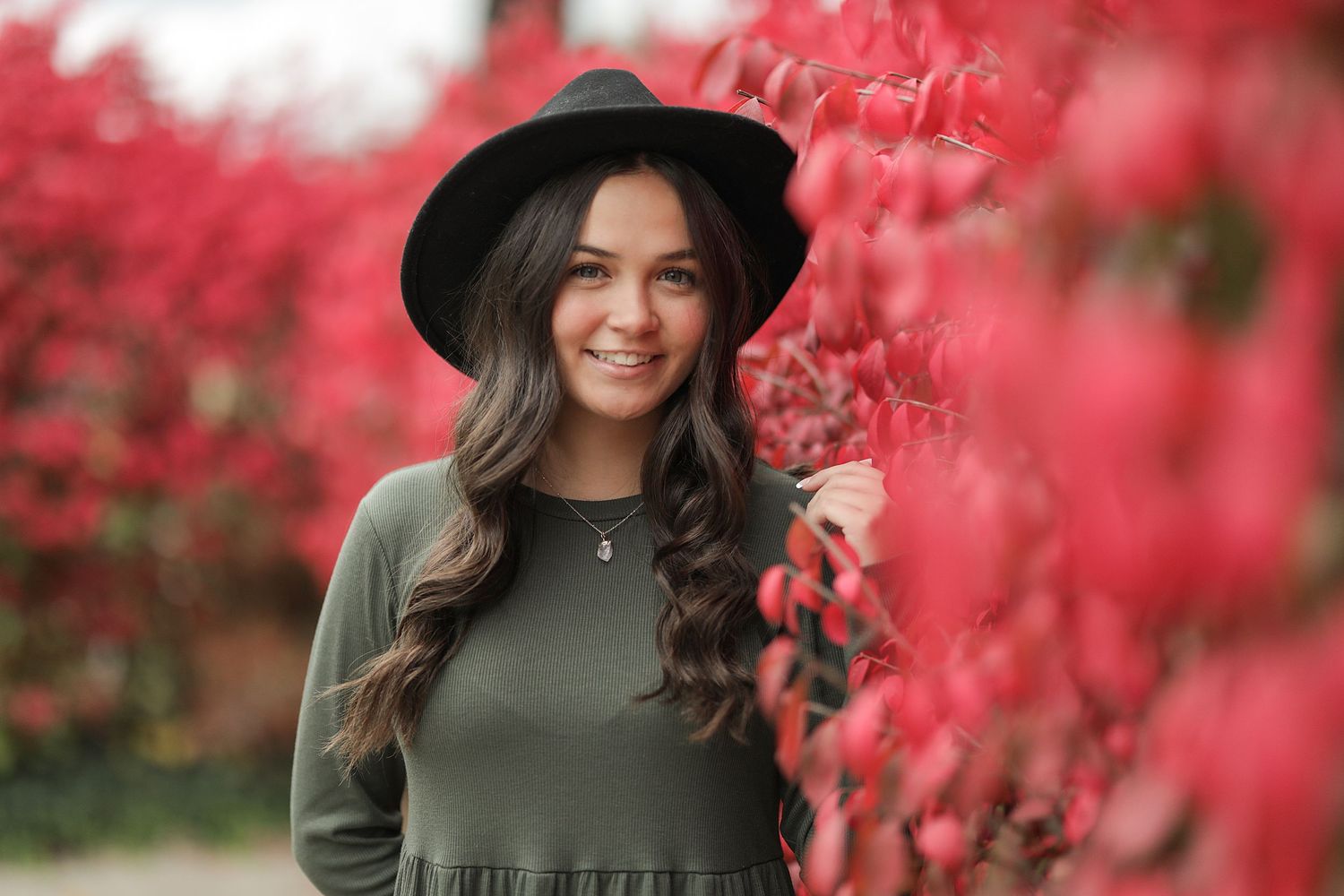 A person in an olive green sweater and black hat poses outdoors among bright red autumn foliage.