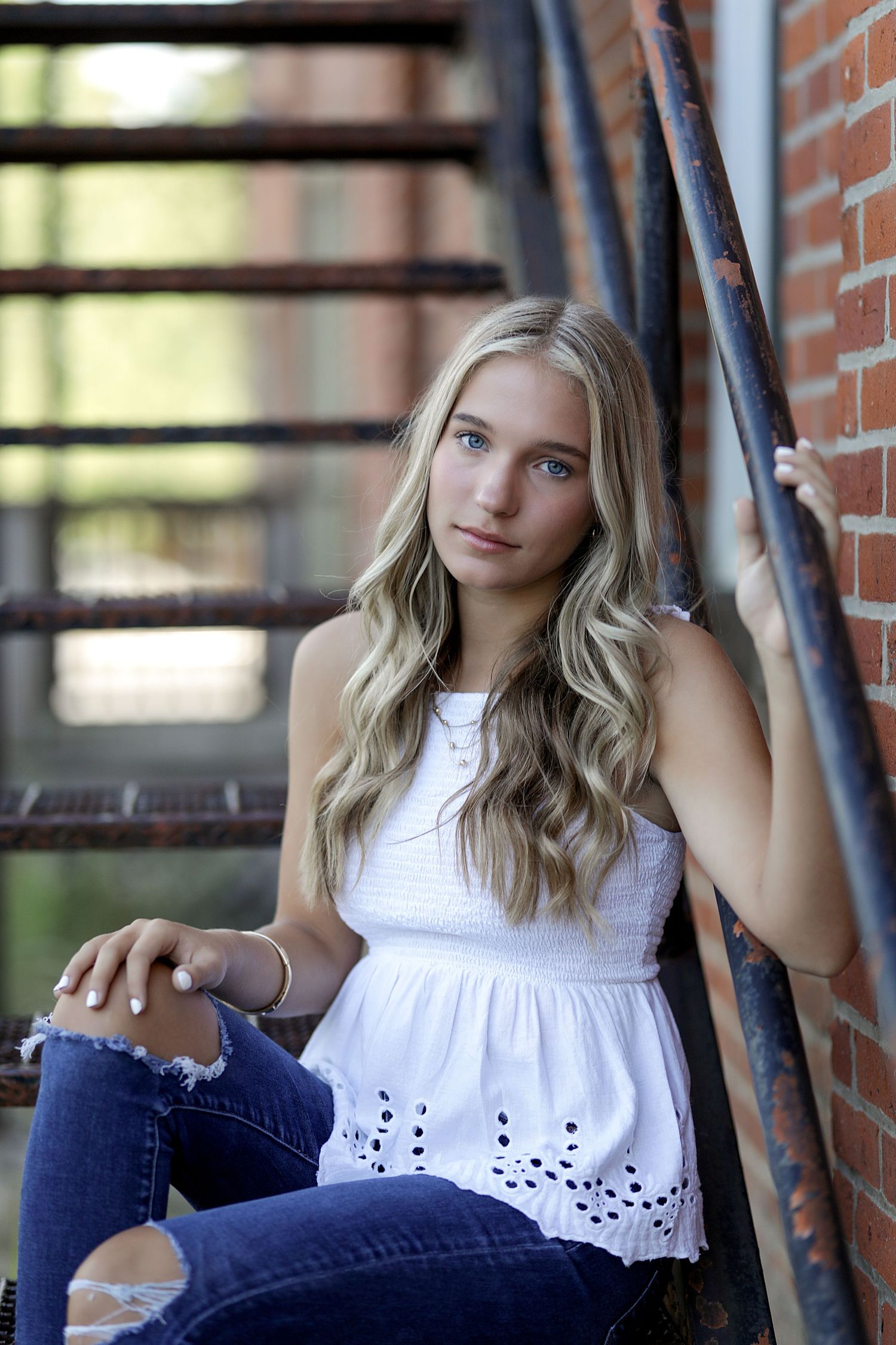 Young person in white sleeveless top and jeans poses casually against brick wall in outdoor portrait setting.