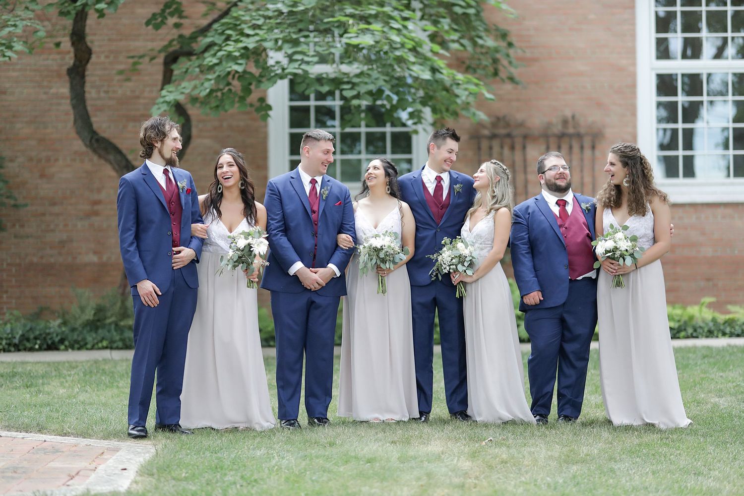Wedding party in navy suits and champagne dresses poses outdoors in front of a brick building.
