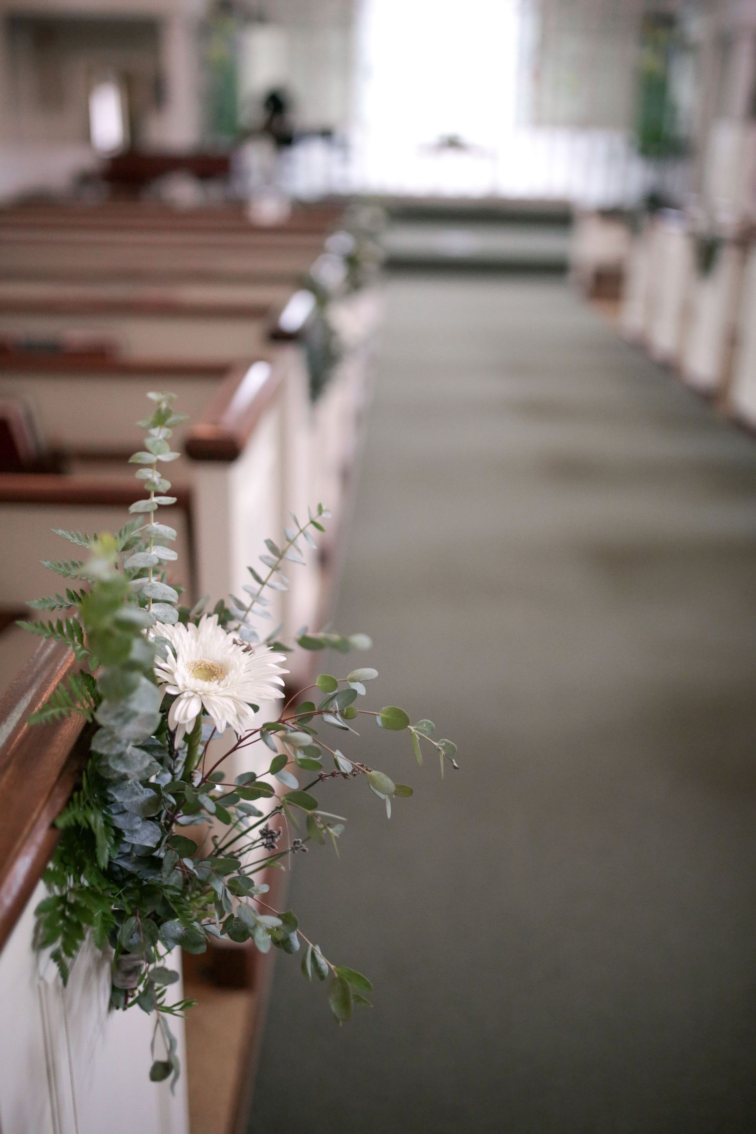 A white daisy and eucalyptus floral arrangement decorates a church pew along an aisle with wooden benches.