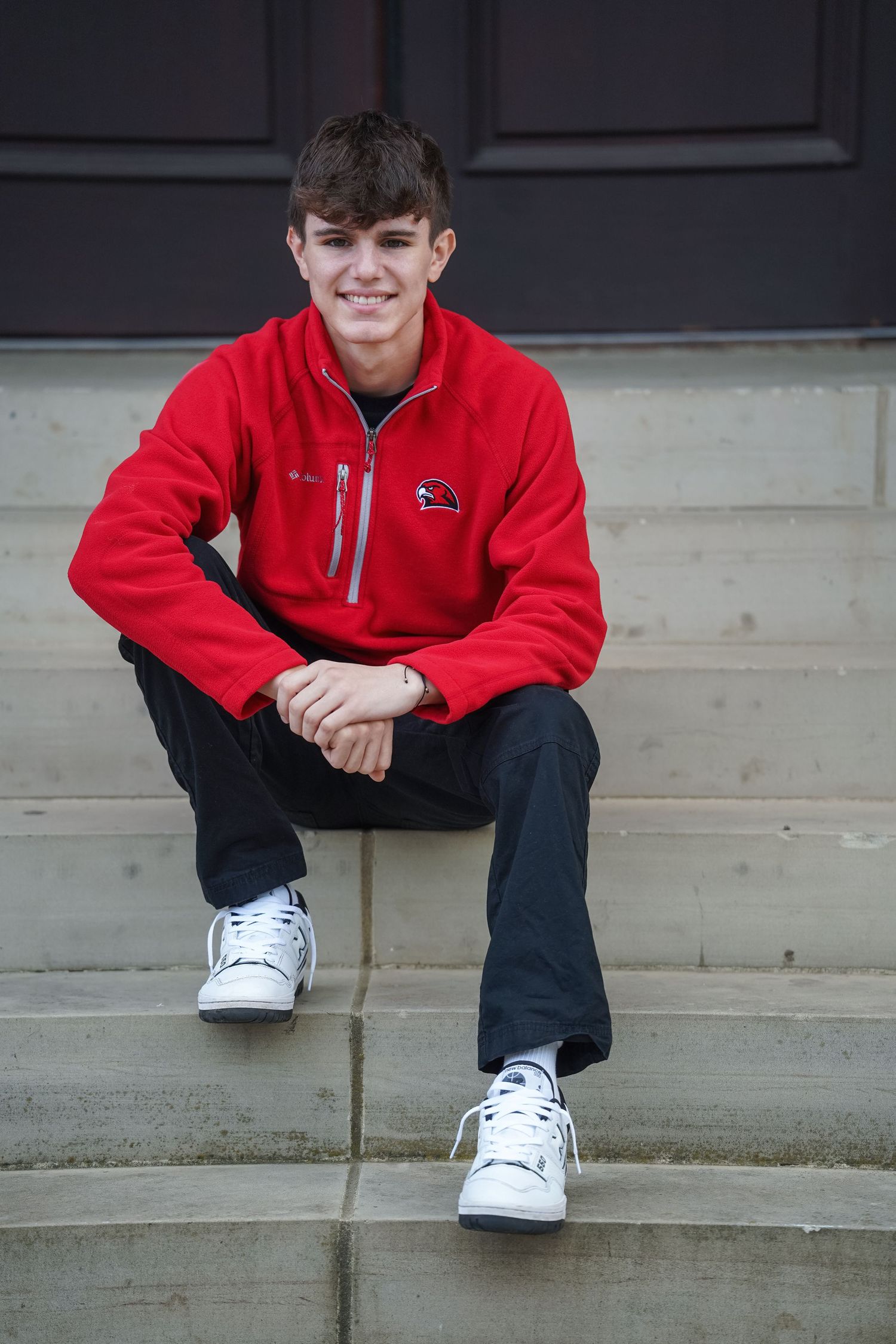 Person in bright red jacket sits casually on concrete steps wearing white sneakers and dark pants.