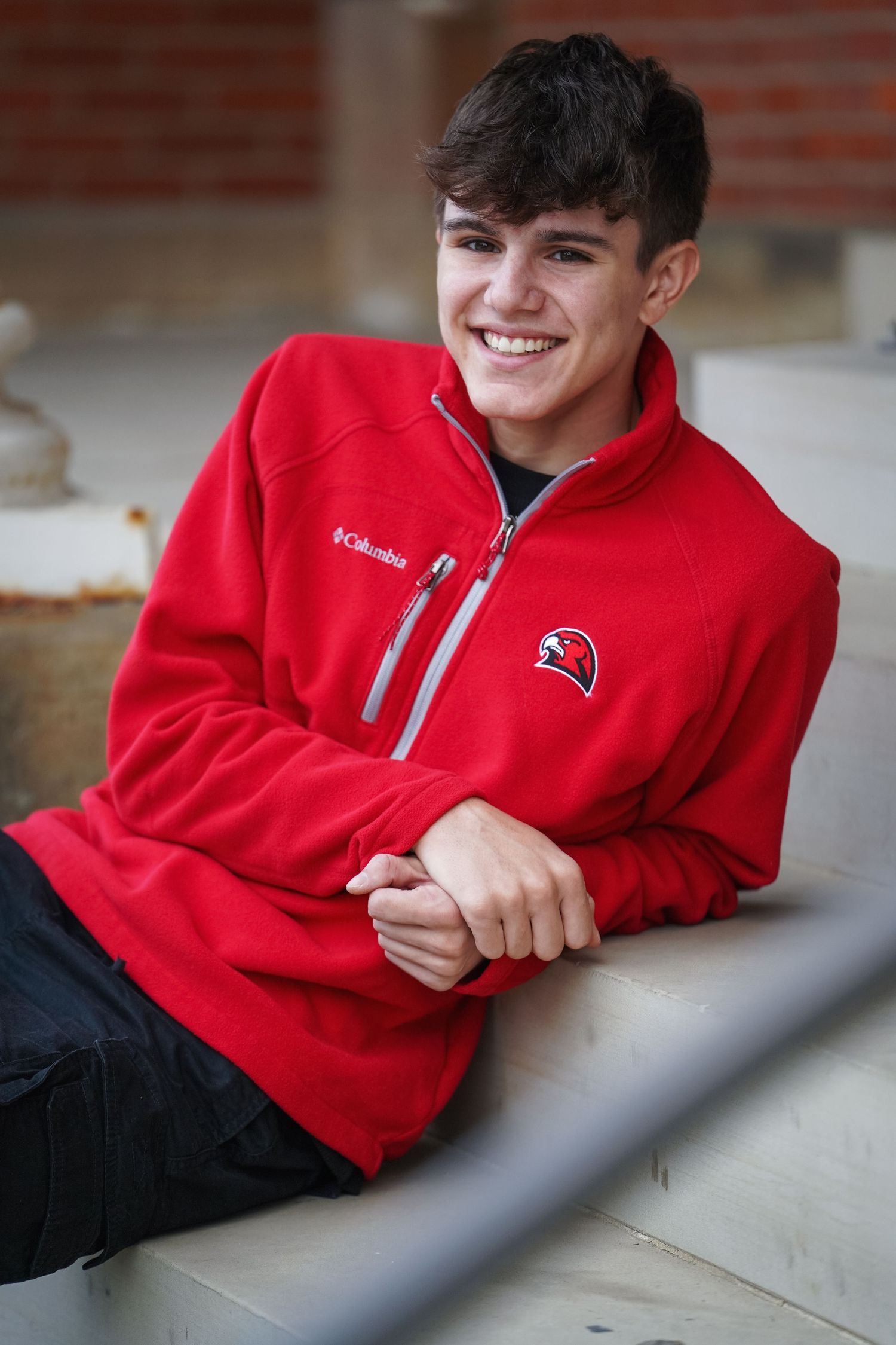A person wearing a bright red Columbia fleece jacket with a team logo sits casually on concrete steps.