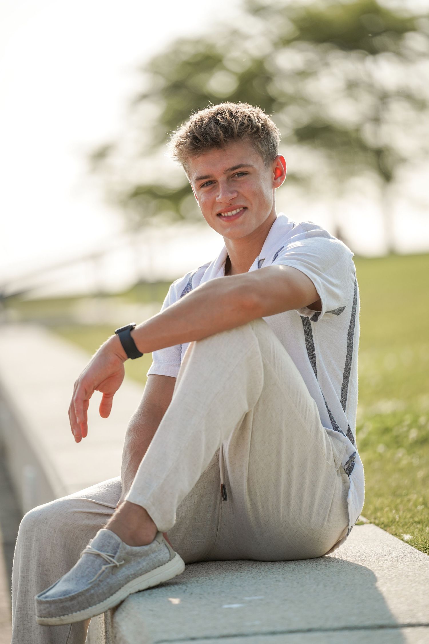 Young person in casual attire sitting outdoors on concrete during golden hour in a relaxed summer pose.